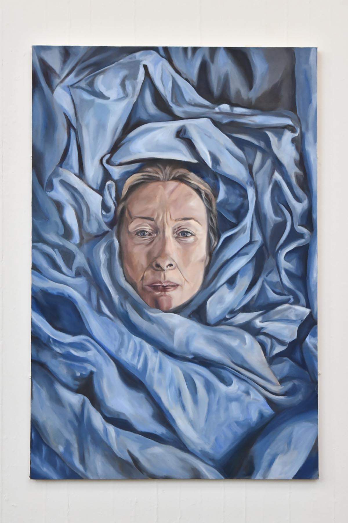 A painting mounted on a white wall of a gallery space. The painting is of a woman's portrait wrapped in blue fabric, her gaze stares towards the viewer blankly.