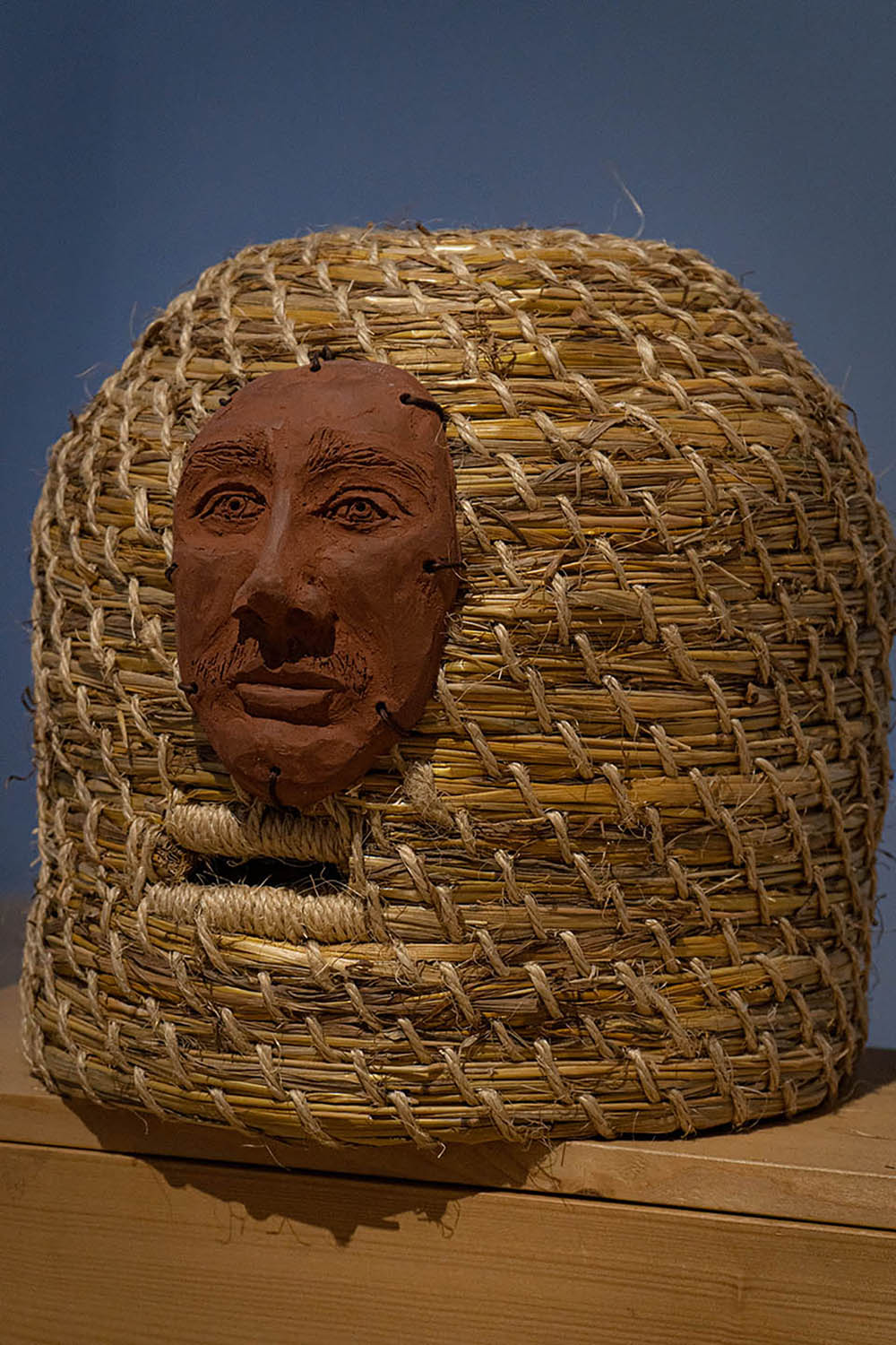 An Image of a Bee Hive/Skep, with a ceramic terracotta male portrait attached to the front, using leather thread.