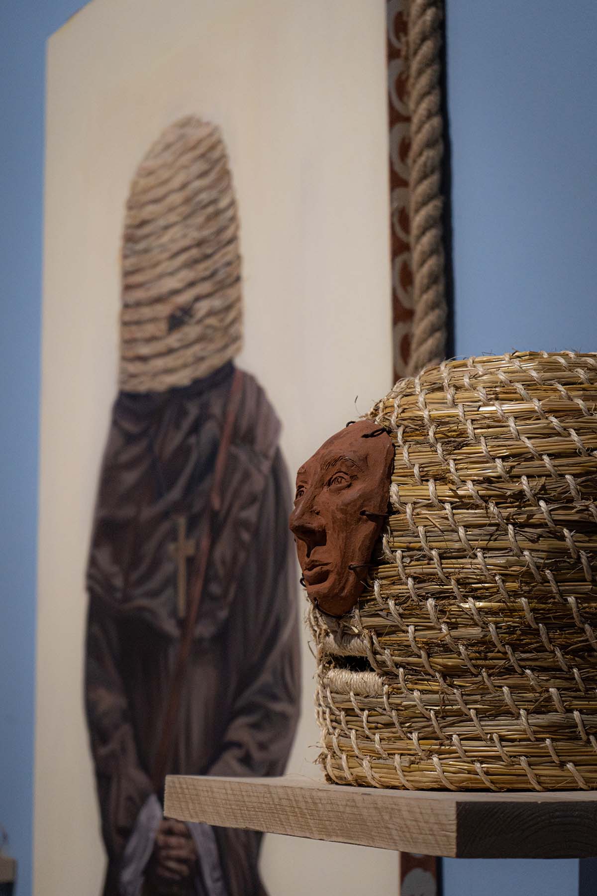 in the foreground there is a beehive sitting on the shelf, a ceramic mask has been attached to the front. In the background there is a monk painting.