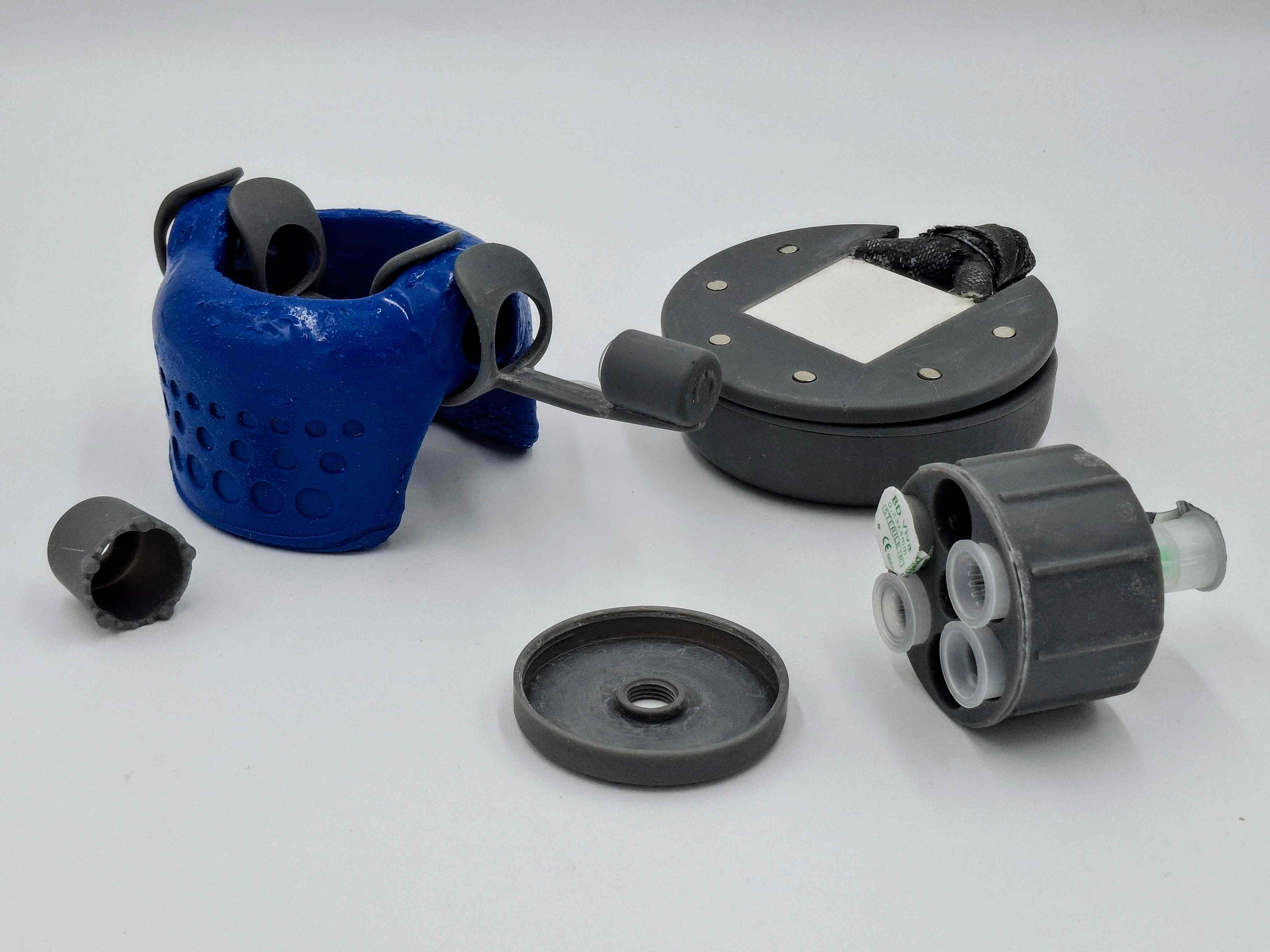 The image shows a blue silicone curved device made of gray plastic component, surrounded by various gray plastic accessories including circular containers, cylindrical pieces, and patterned components. A circular gray device with white center elements sits in the background.