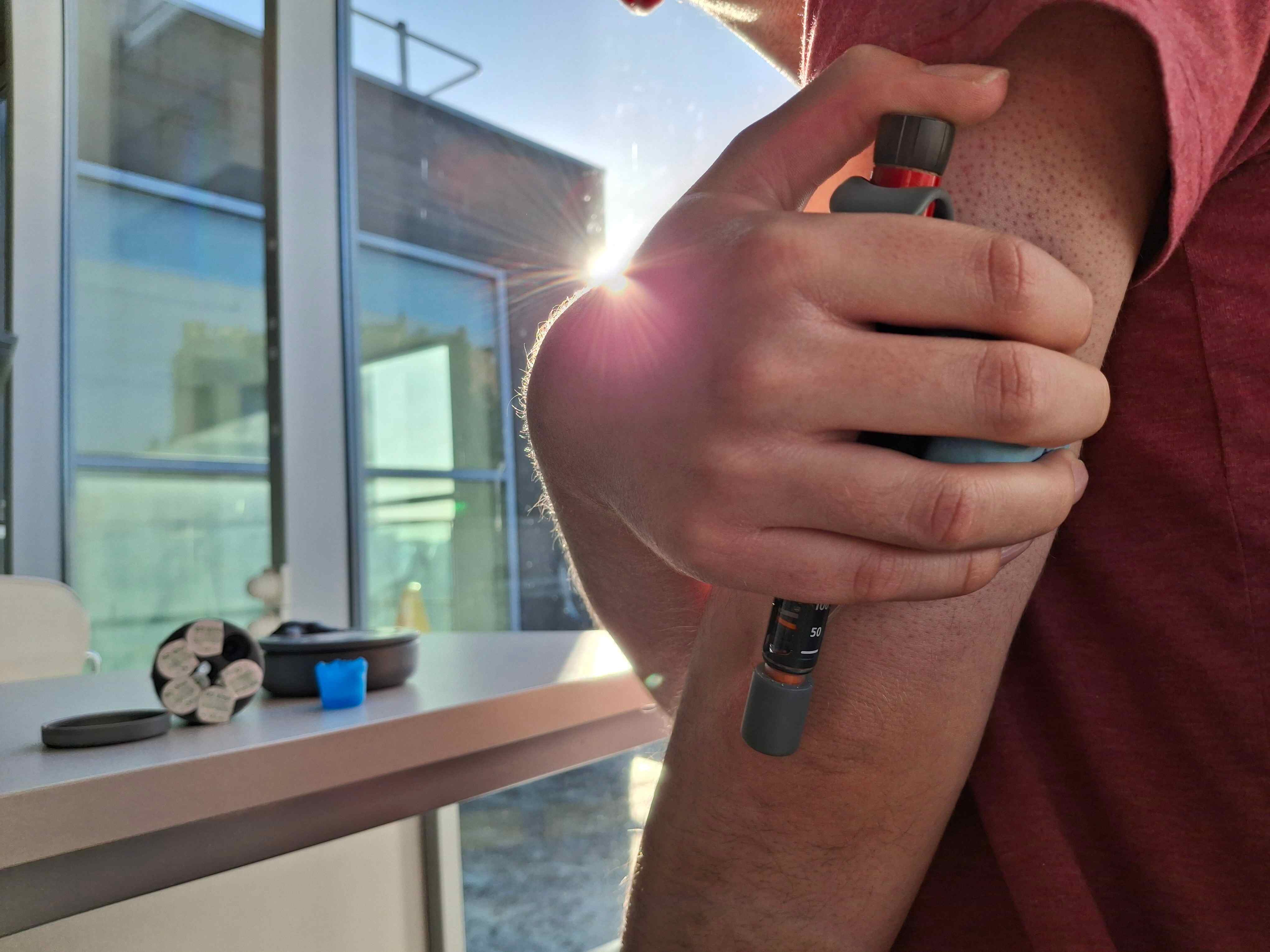 The image shows a close up of someone holding the Hylow injector with a hugging motion, ready to inject whilst the rest of the designs are on the table at the background of the image.