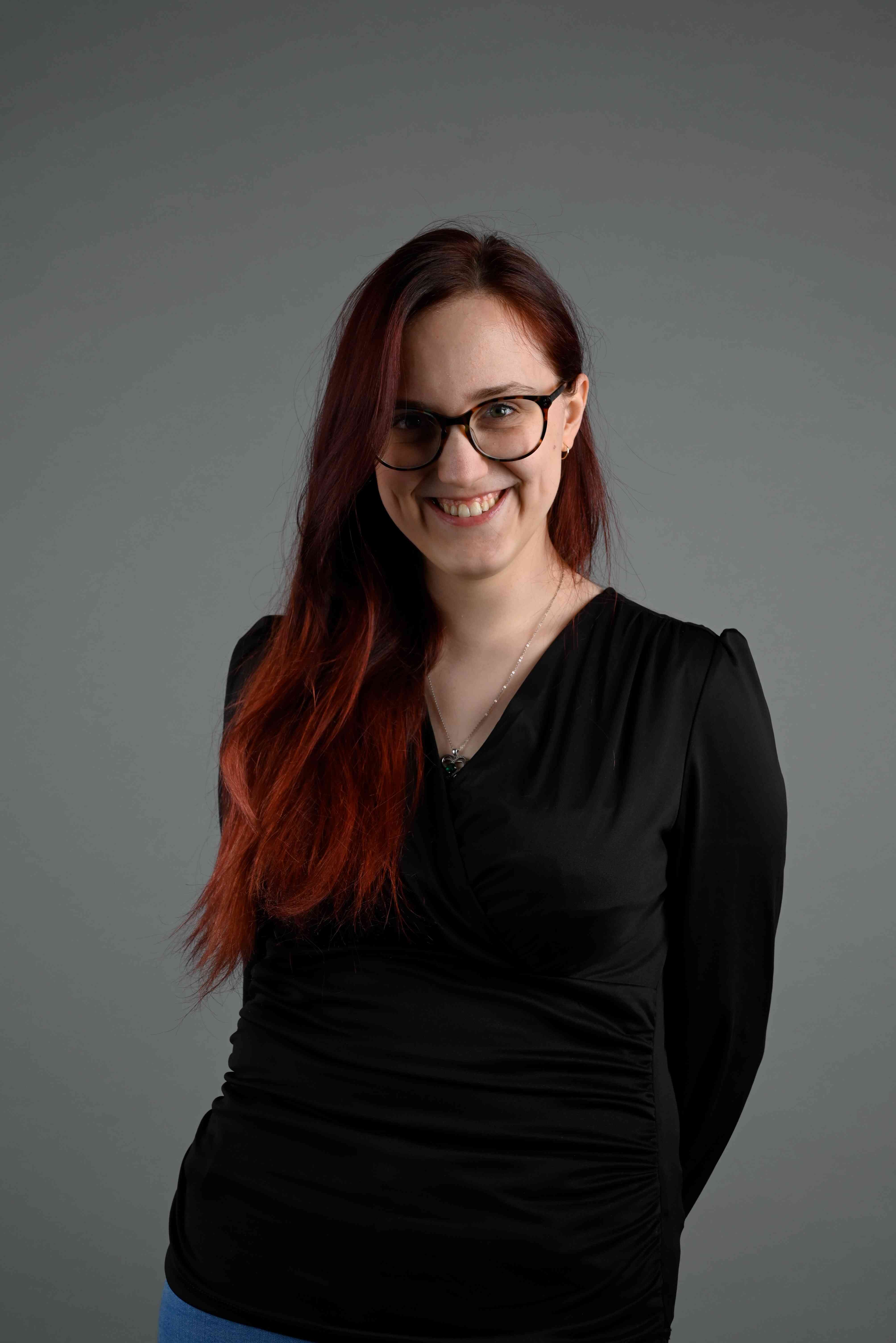 Viktoria Simon with red hair and black smart long sleeved top on a grey background.