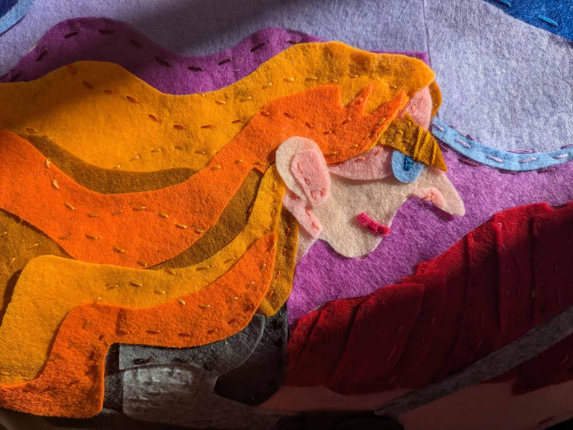 A zoomed in felt applique image of a woman with flowing orange hair riding on a unicorn. She wields a needle as if it is a sword.