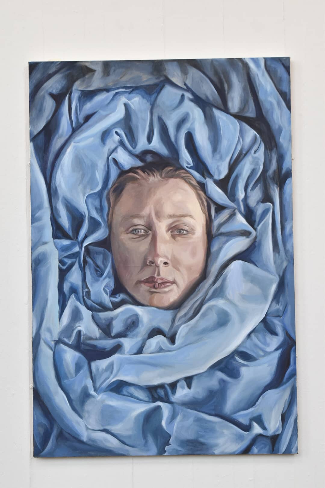 A painting mounted on a white wall of a gallery space. The painting is of a woman's portrait wrapped in blue fabric, her gaze stares towards the viewer blankly.