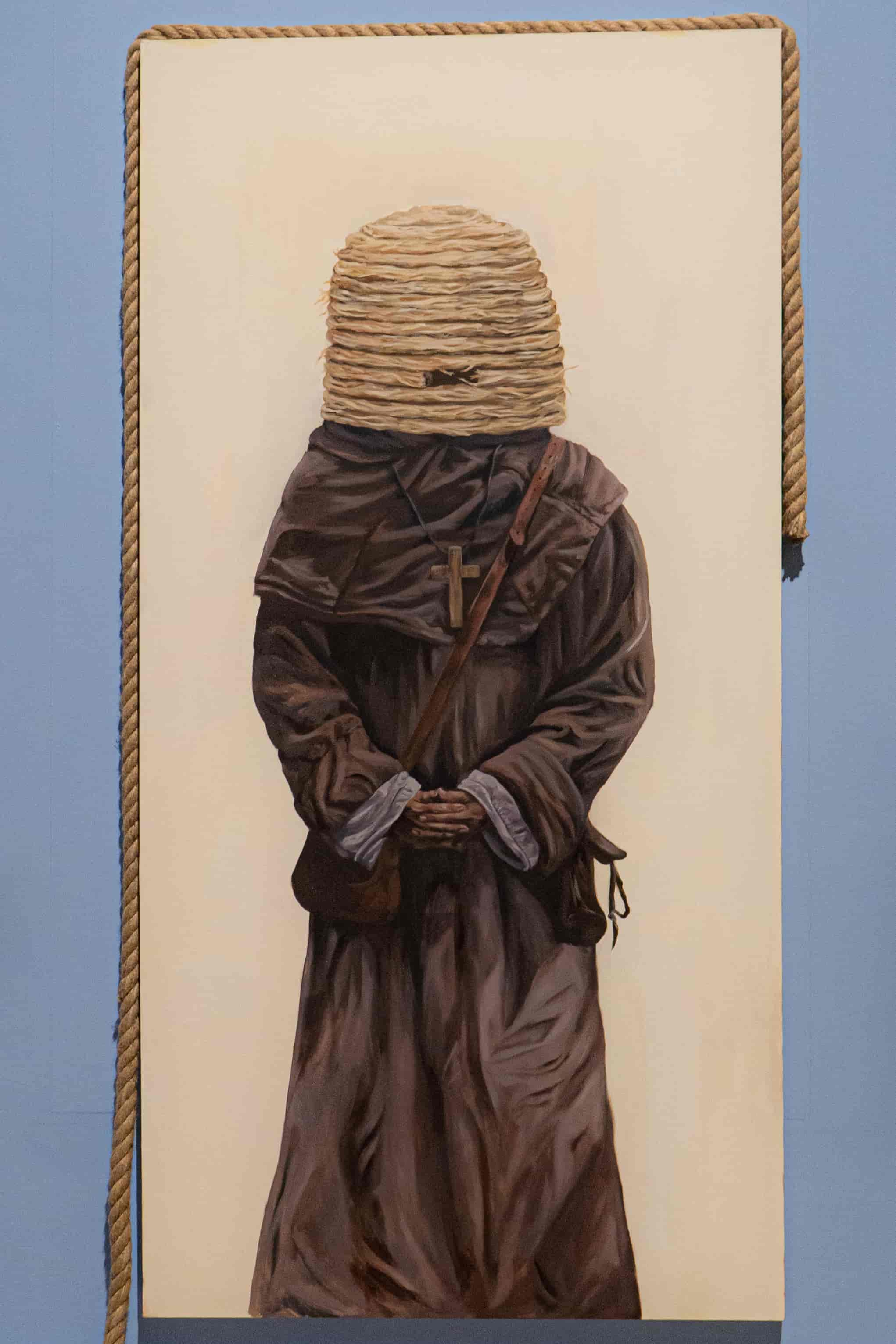 An image displaying a painting on a blue wall. The painting is that of a monk figure with a bee hive over his face. There is also a rope which dangles asymmetrically around the work.
