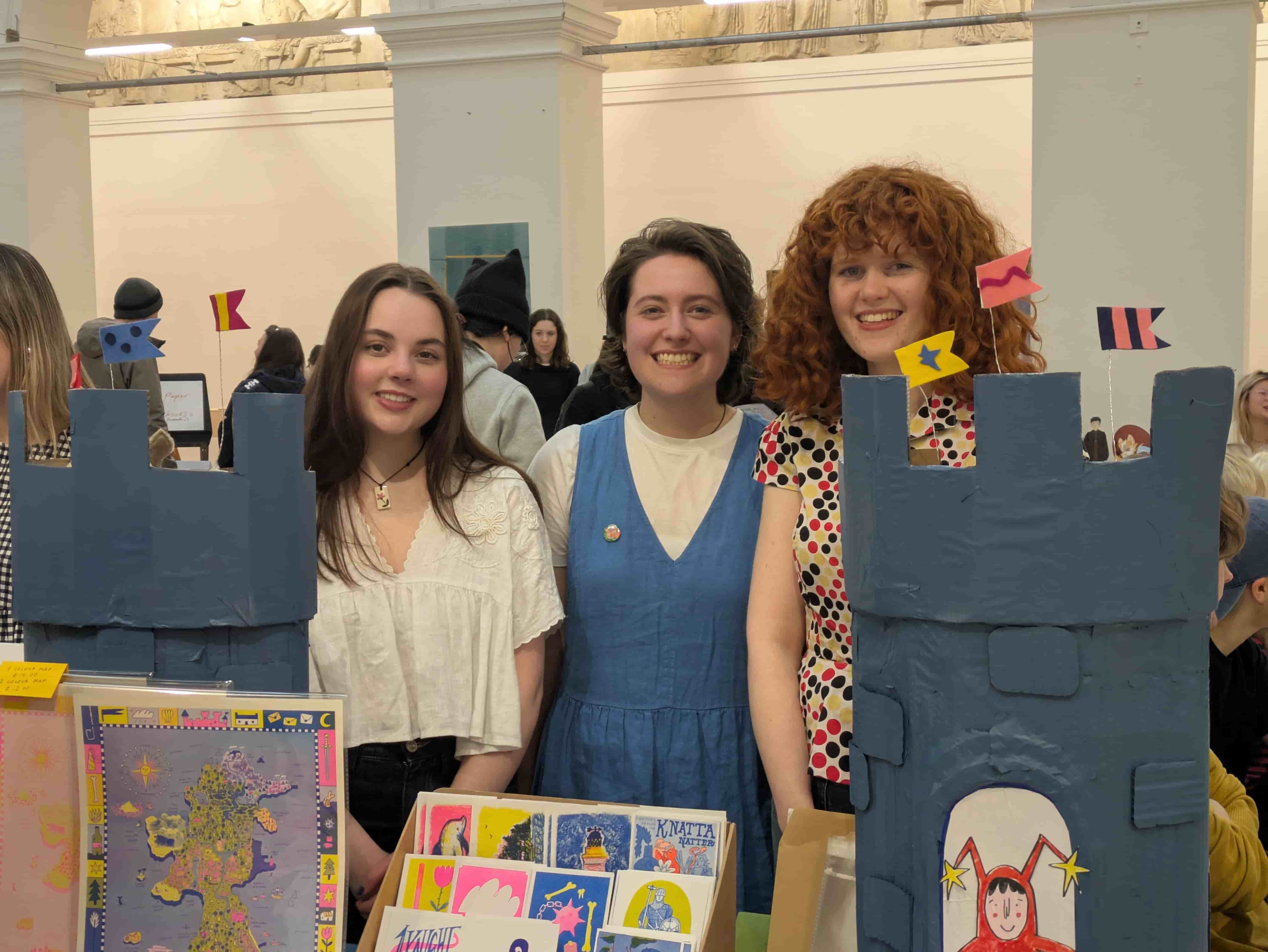 Three people stand smiling behind an artist fair stall