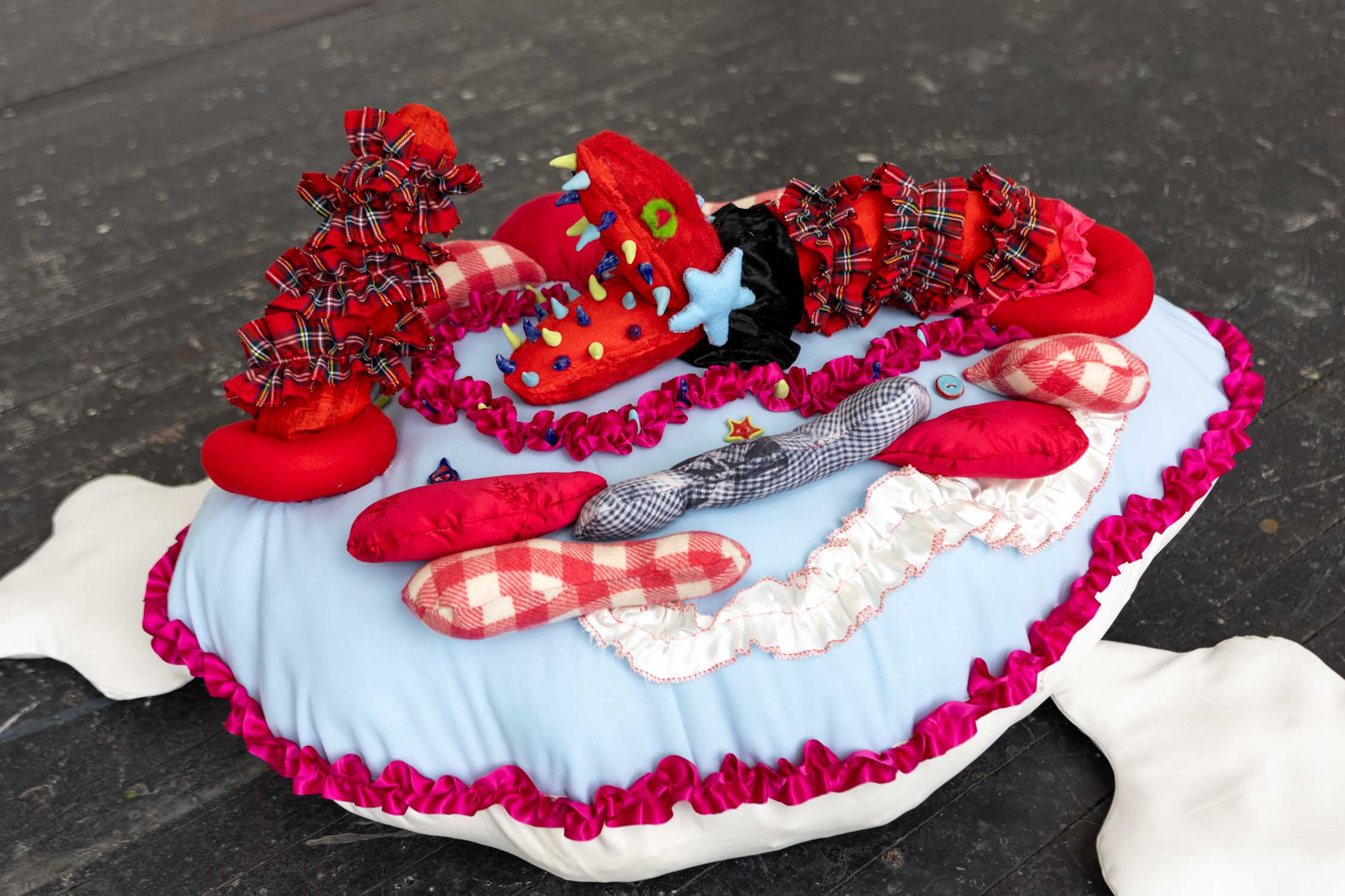 Soft sculpture of a stuffed ionic shape with a red snake protruding out of it, trying to eat its own tail. The sculpture is covered in ruffled ribbon and some patterned fabric, such as gingham and tartan. 