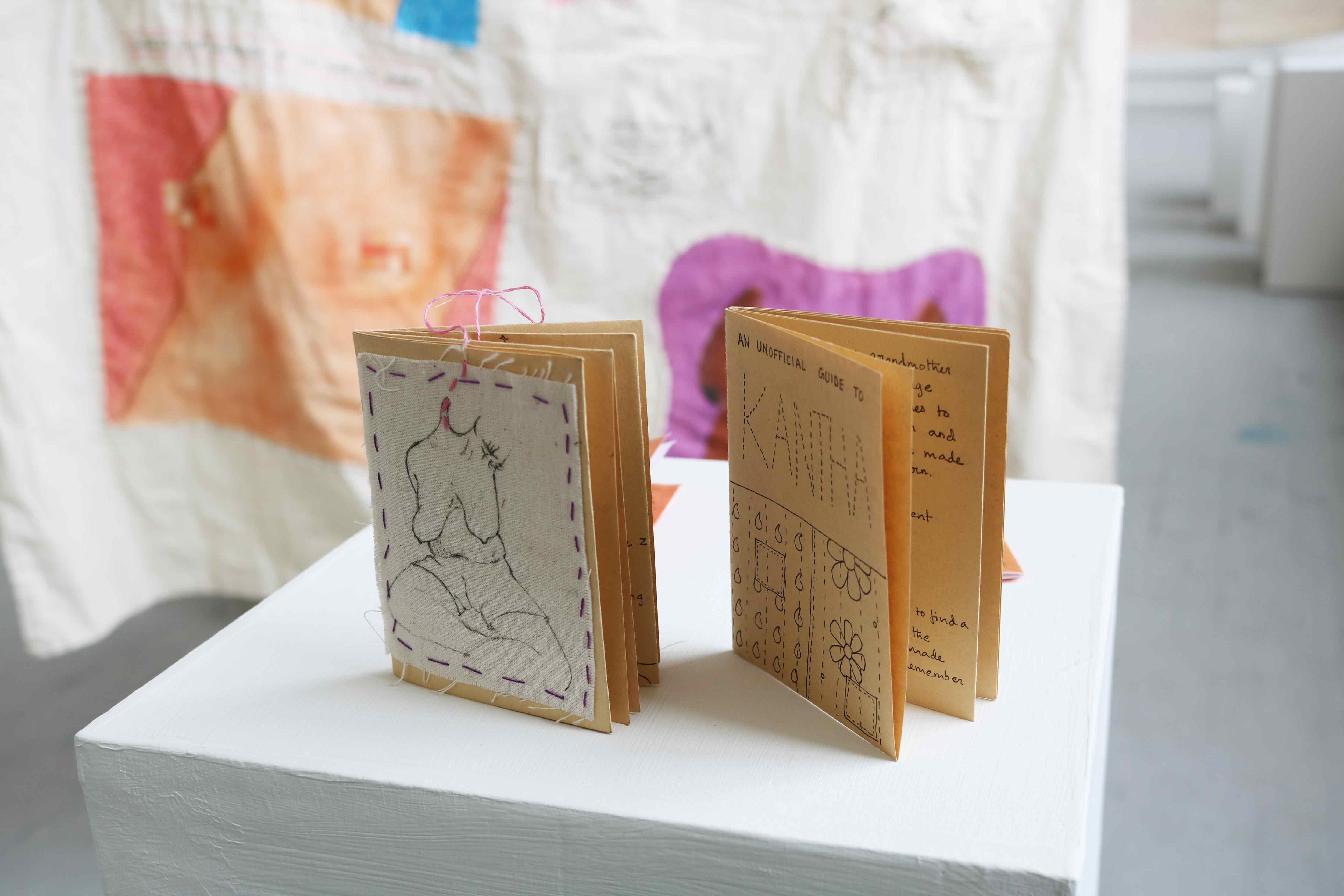 Couple of Zines on a plinth, One zine has a cover where a piece of fabric with an illustration has been sewn onto the paper, it is a line illustration of a topless body where the neck turns into a a thread functioning as a bookmark. The other Zine has illustrations and writing with the over saying 'KANTHA'