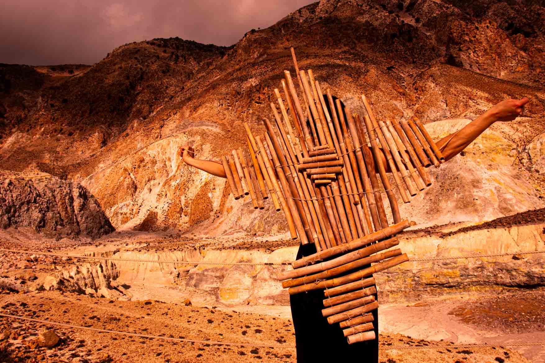 Dora Zoi standing in the rocky volcanic landscape of Nisyros, Greece, wearing a bamboo sculpture on her back during an artistic research trip for the Paolozzi Travel Award. Photograph by Anton Vasiliadis. August 2025.
