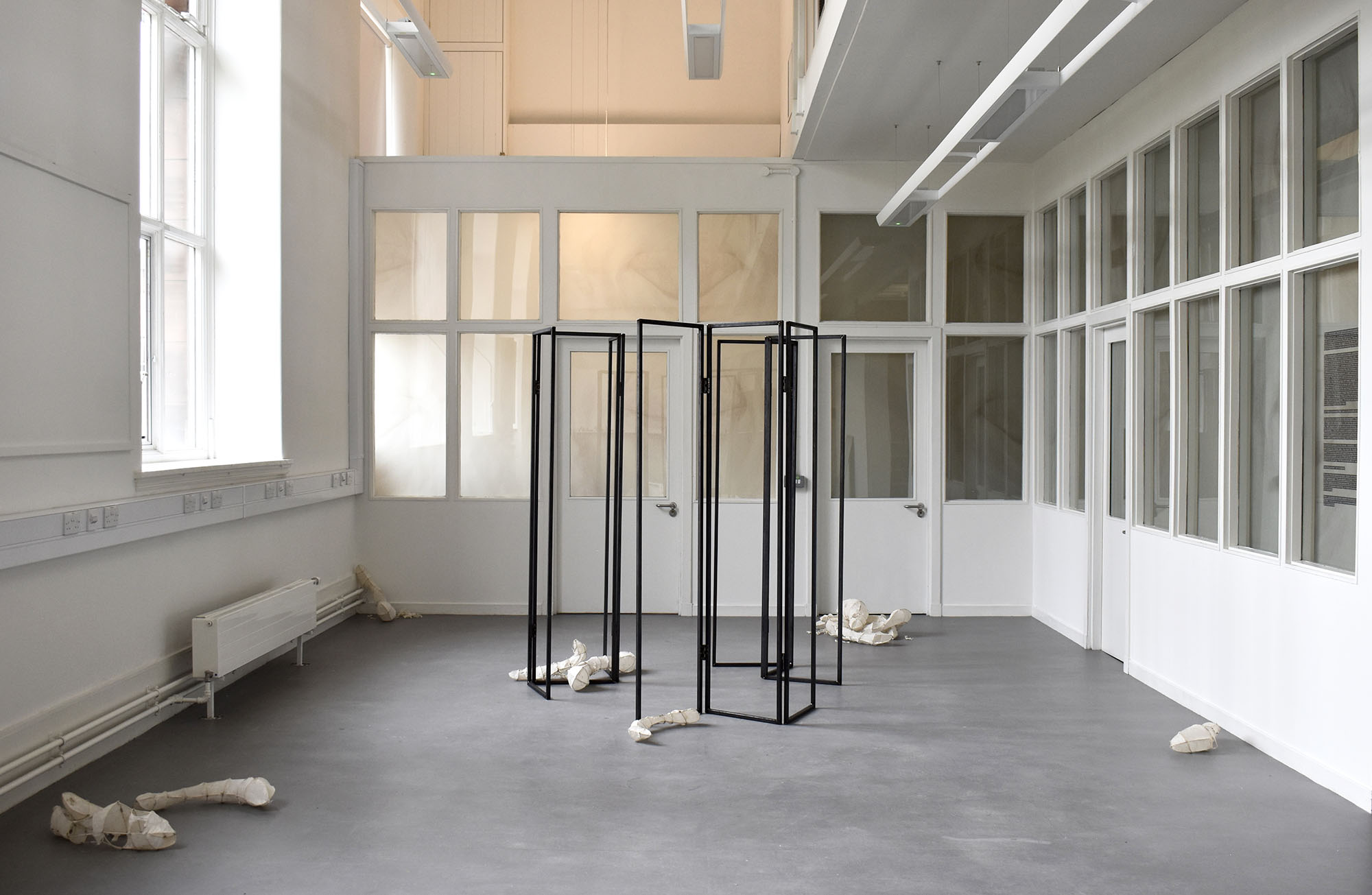 A wide installation view of two windowed walls. Each window has tracing paper prints with closed eyes covering them from the inside. Three hollow steel dressing screens and a variety of rice paper sculptures occupy the floor space before the windows.
