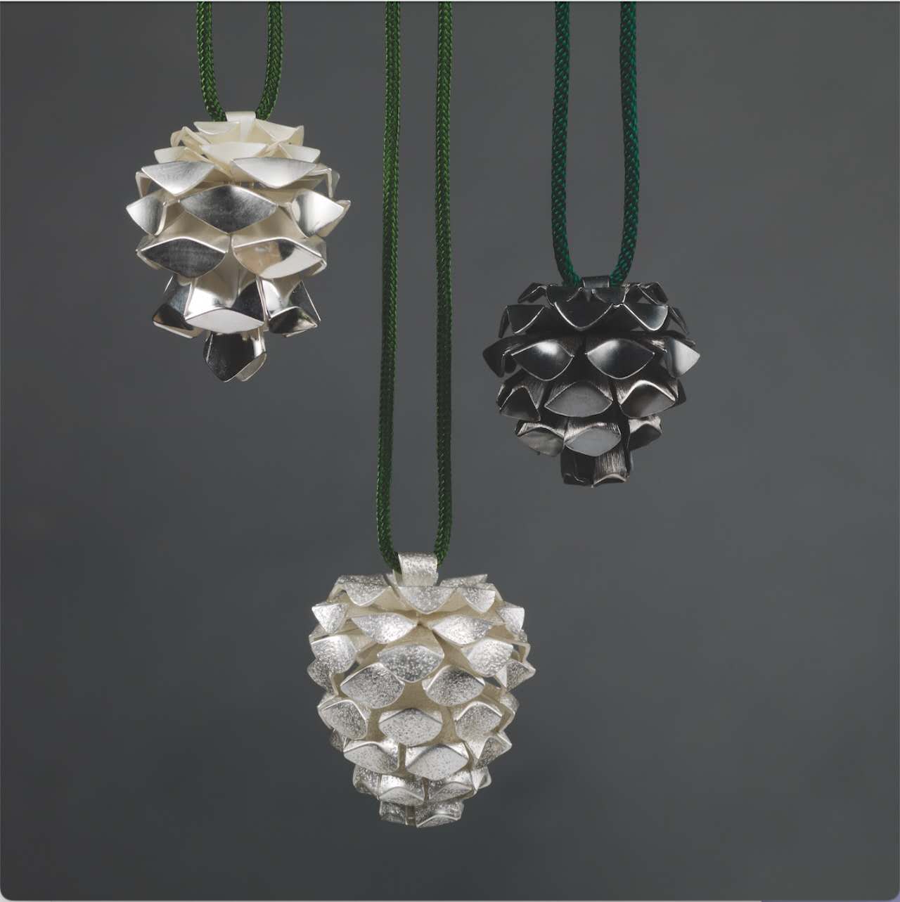 They are three small pinecone-inspired structures. On the left is a silver pinecone bell necklace with wide layered scales and a gradient colour effect. In the centre is an elongated silver pinecone form with textured surfaces. On the right is a small black-and-silver pinecone necklace featuring interlocking layered elements.