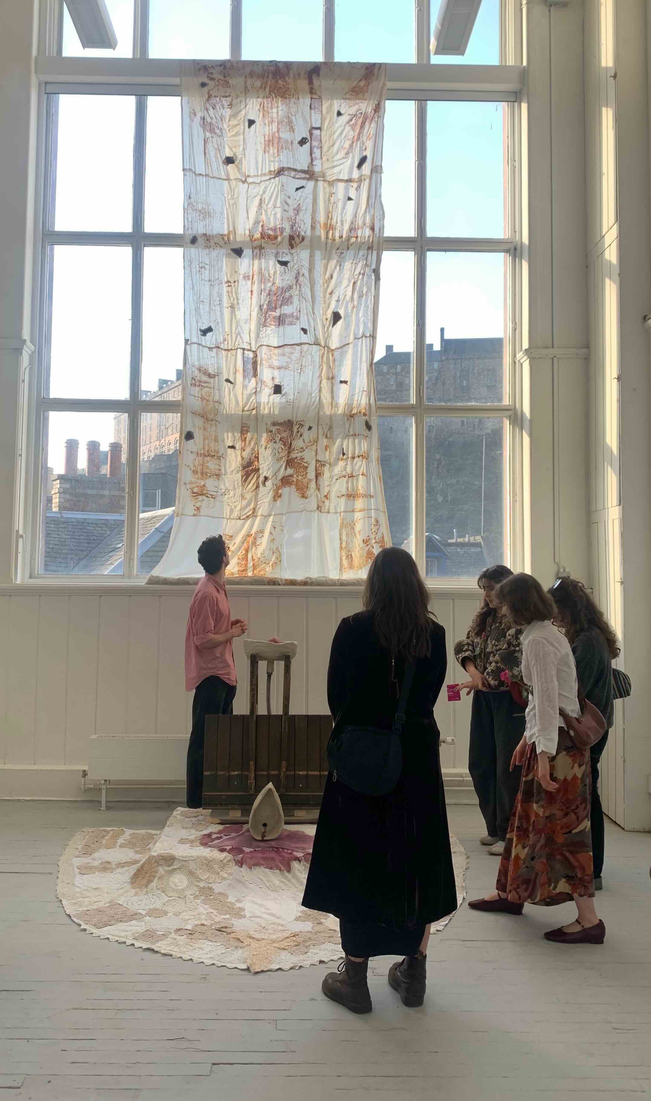 Group of people gathered around a sculptural installation featuring textiles on the window and floor and a wooden and ceramic centrepiece.