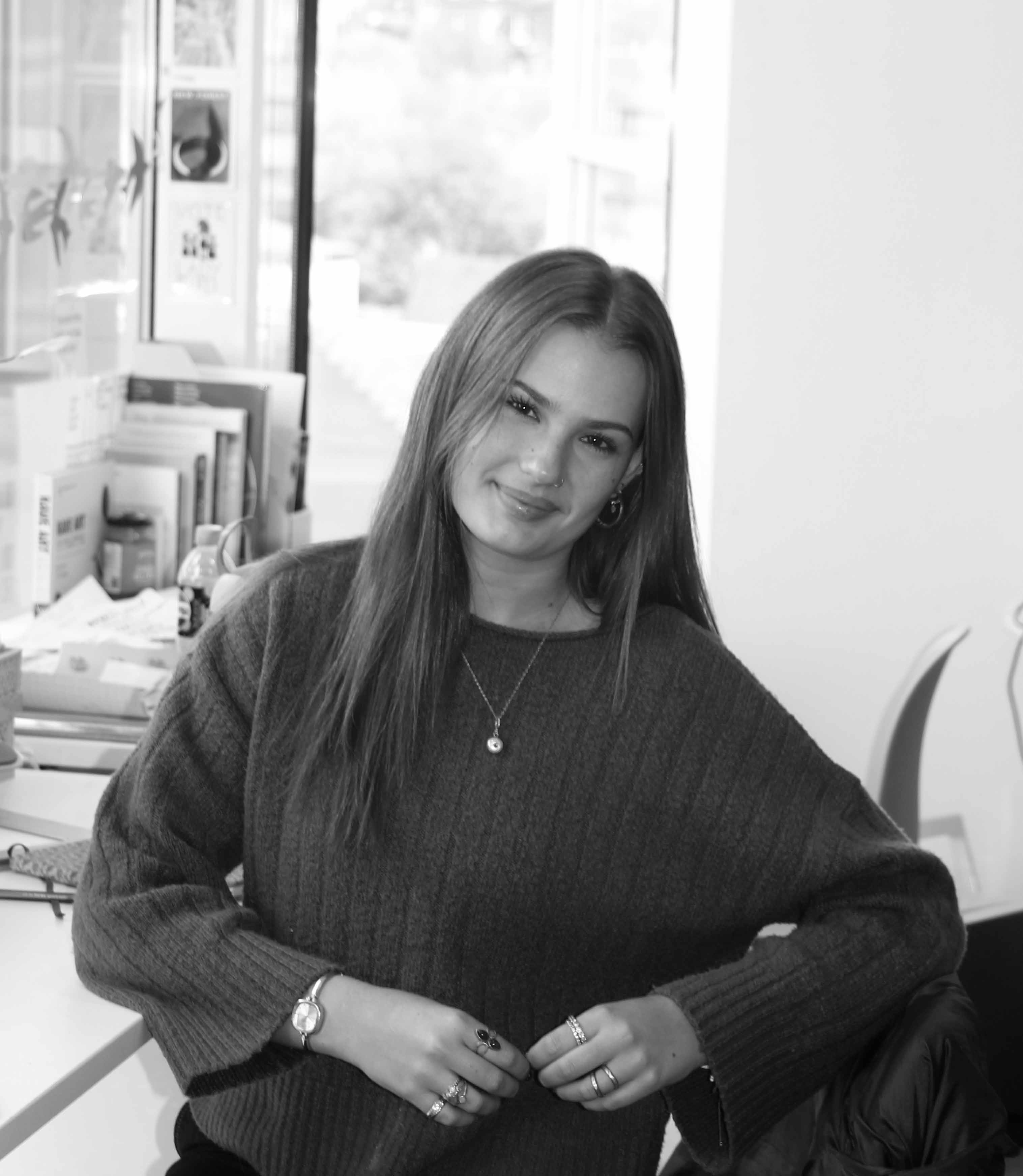 Black and white portrait of a young woman sitting at a desk in a studio or workspace. She is smiling towards the camera and wearing a dark knitted jumper, jewellery, and a necklace. Books, papers, and creative materials are visible in the background near a large window.