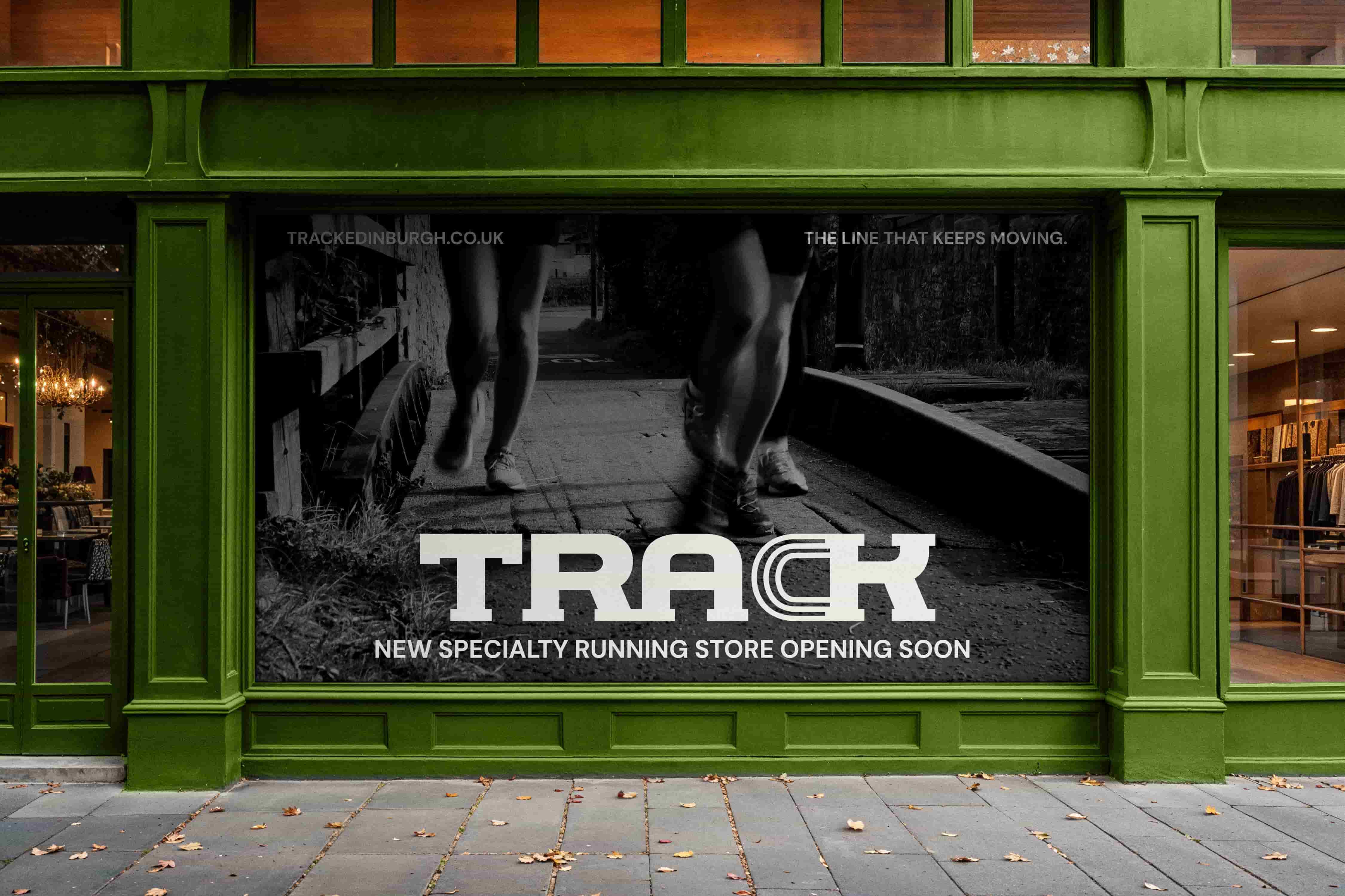 Black and white storefront poster for TRACK, a new specialty running store. The poster shows two runners moving along a path, with the TRACK logo centred in bold white text and the wording ‘NEW SPECIALTY RUNNING STORE OPENING SOON’ underneath. Smaller text at the top reads ‘trackedinburgh.co.uk’ and ‘THE LINE THAT KEEPS MOVING.’ The poster is displayed across the front window of a dark green shop exterior.