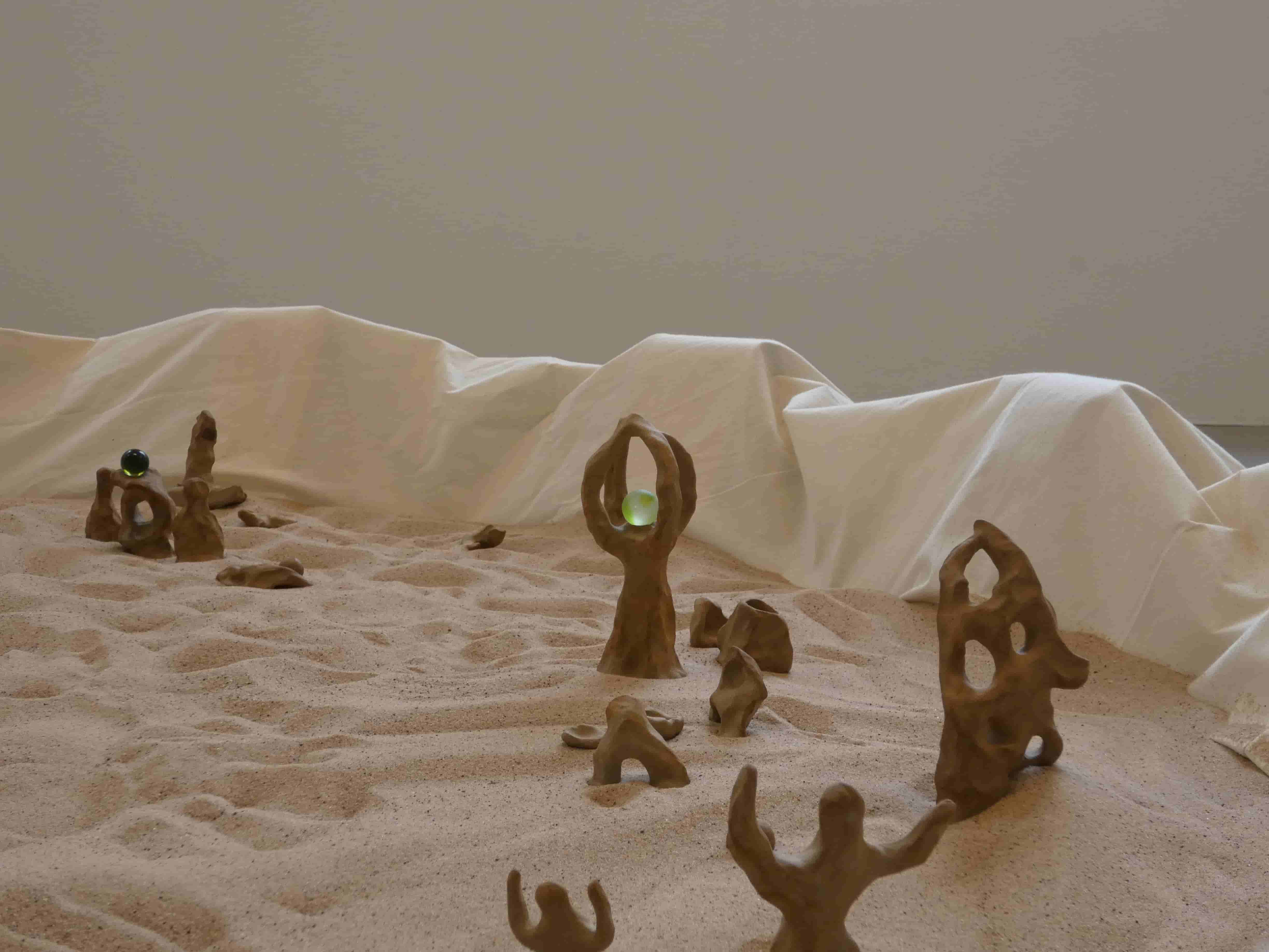 Abstract sculptural figures and two glass marbles sit in sand, fabric wall of the pit drapes behind  