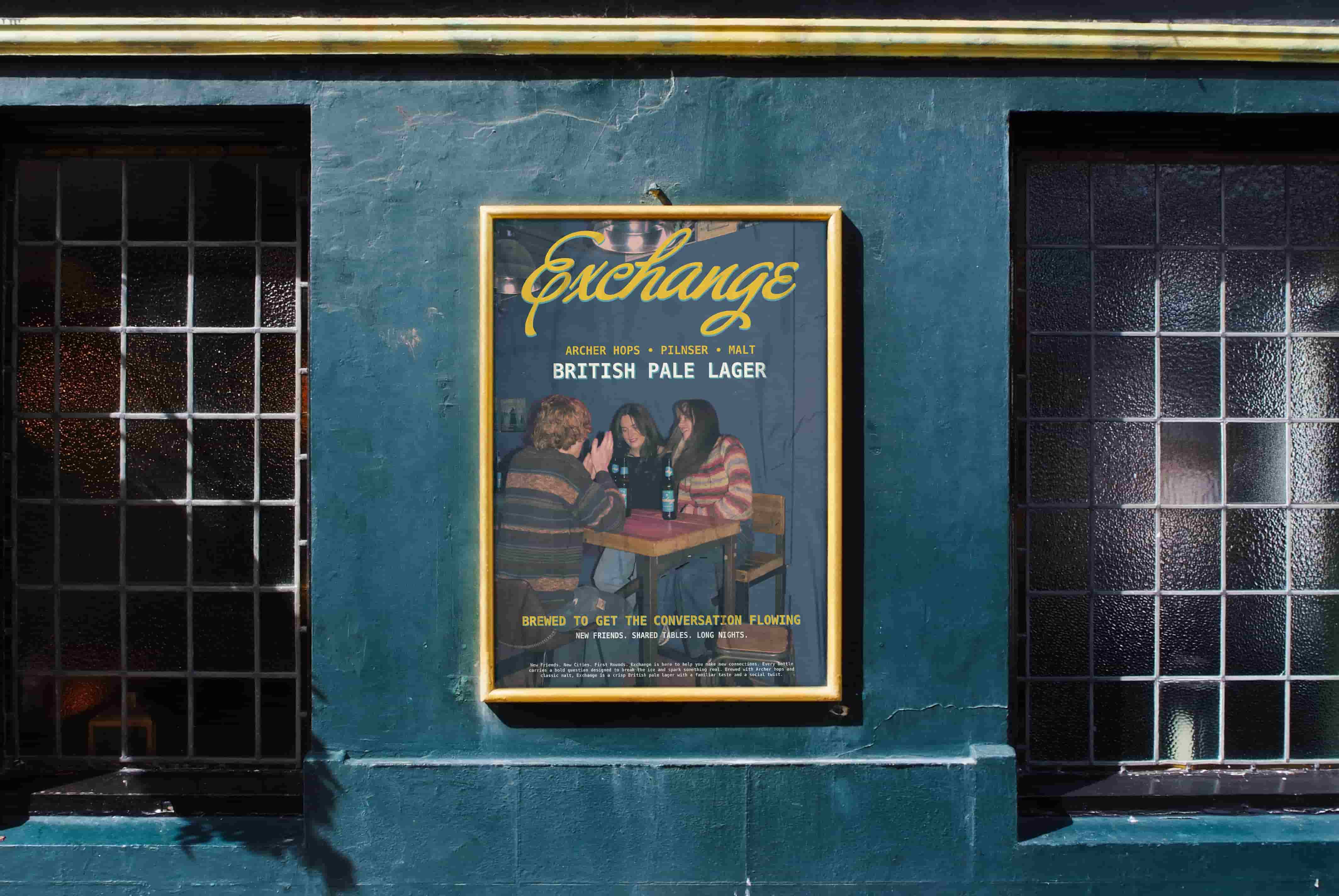 Framed poster for ‘Exchange’ British Pale Lager displayed on a teal pub wall between two dark, gridded windows. The poster shows three people sitting around a table with beer bottles, with text promoting conversation, new friends, shared tables and long nights.
