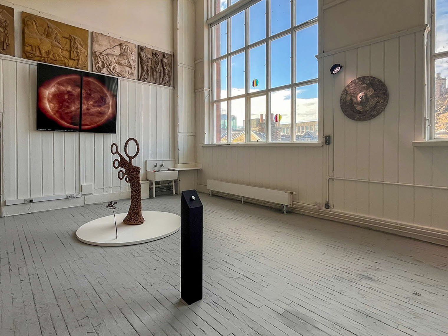 Installation view of an abstract mosaic figure in brown ceramic tiles on a white round plinth, beside a bronze cast of hogweed. Screens display a sun image, and windows feature two circular pieces of fused glass.