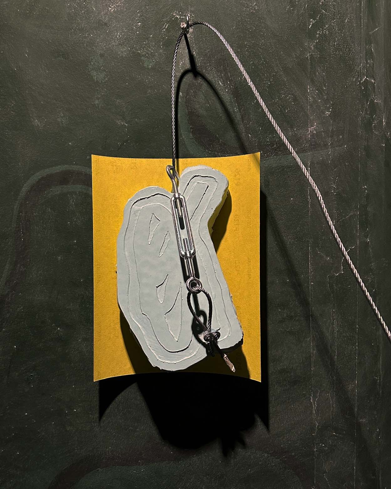 steel wire hanging a blue silicone sculpture, in front of a sheet of yellow sandpaper, on a dark green wall