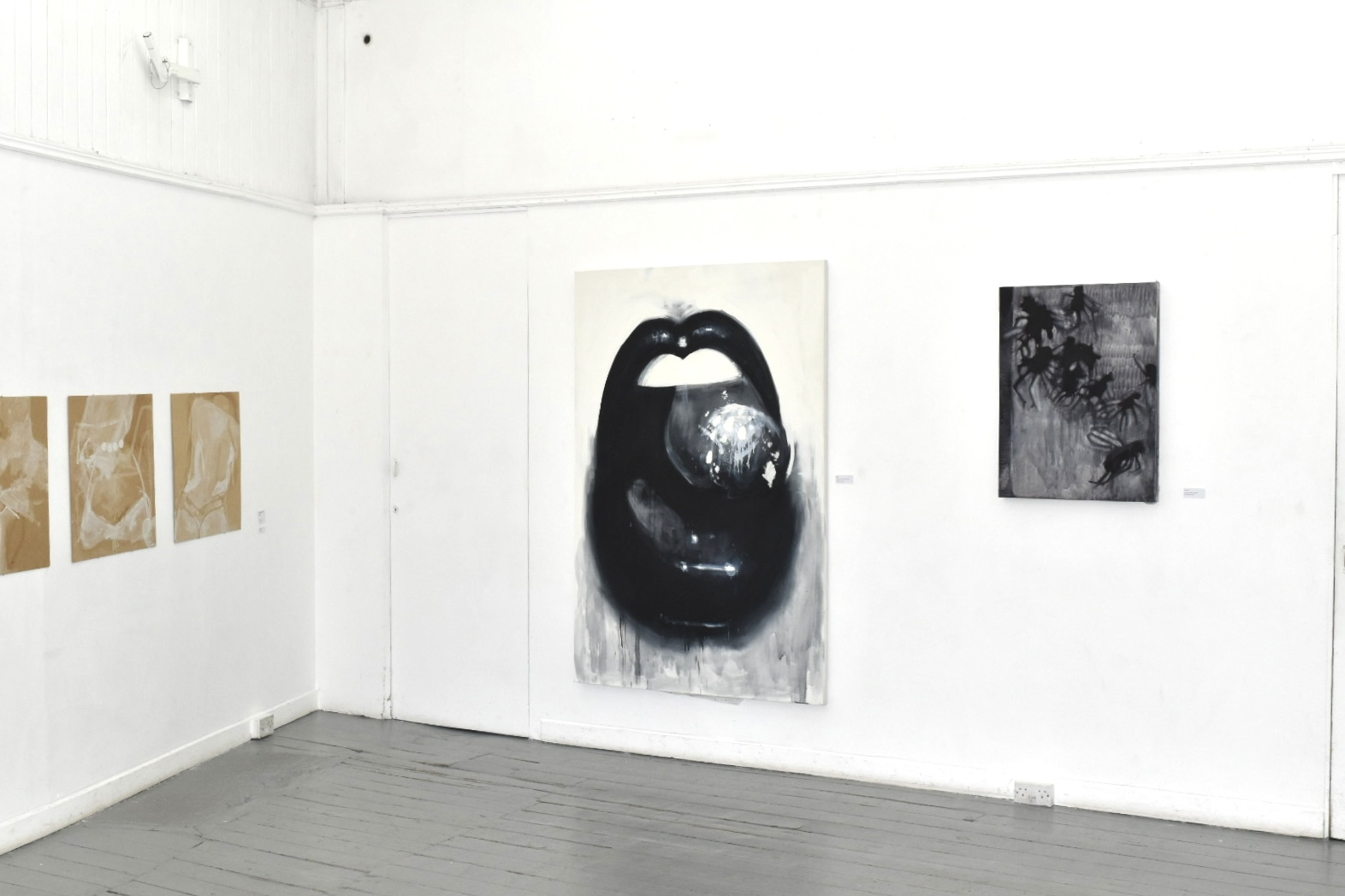 exhibition setting with large black and white paintings and another artists brown and white paintings, big mouth next to dead flies