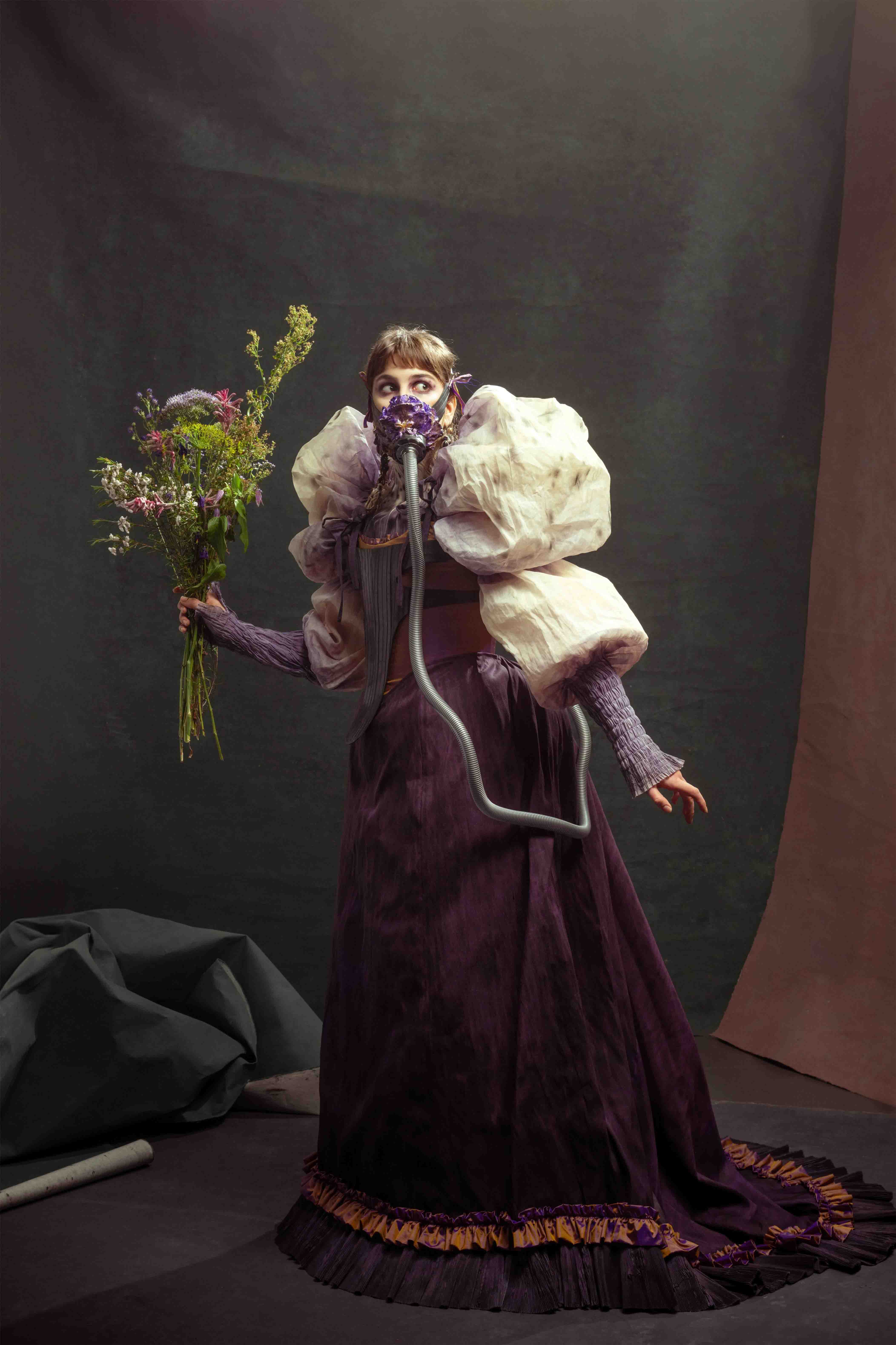 A woman in a purple dress with very large cream puff sleeves. She is wearing an oxygen mask that looks like a purple flower and has a silver tube running from it and into the back of the skirt. She is holding a bunch of flowers.