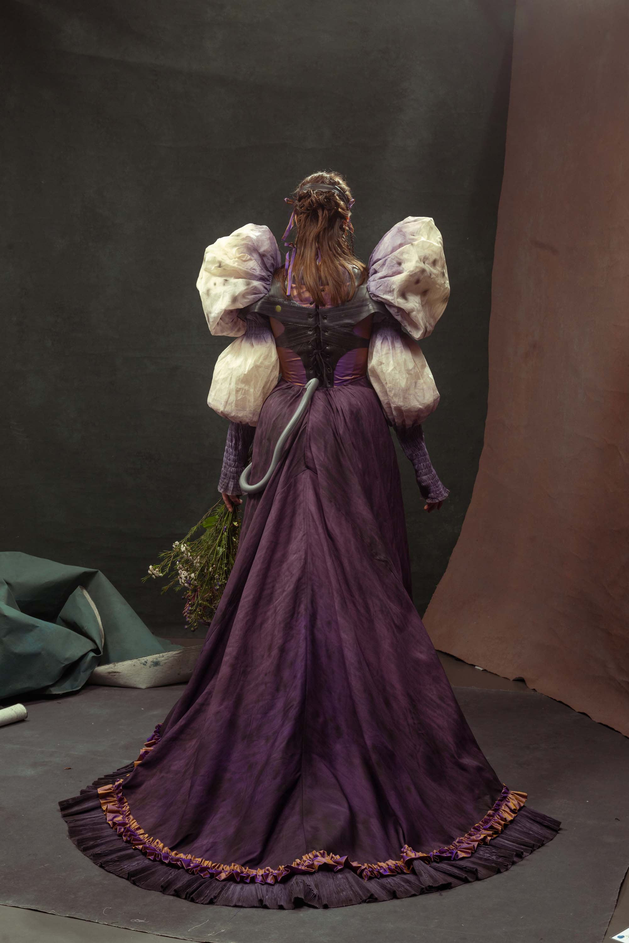 A woman in a purple dress with very large cream puff sleeves. She is wearing an oxygen mask that looks like a purple flower and has a silver tube running from it and into the back of the skirt. She is holding a bunch of flowers.