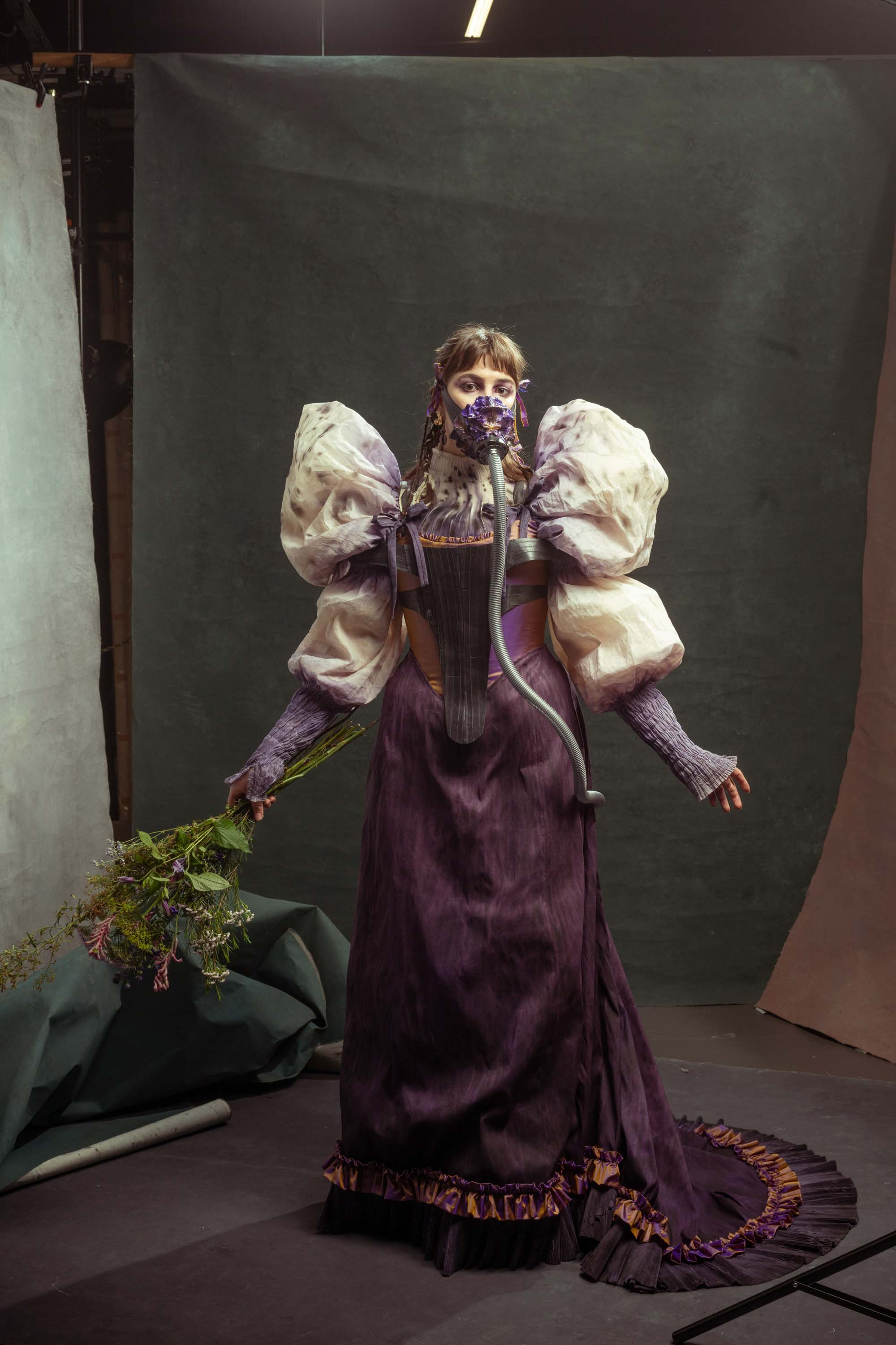 A woman in a purple dress with very large cream puff sleeves. She is wearing an oxygen mask that looks like a purple flower and has a silver tube running from it and into the back of the skirt. She is holding a bunch of flowers.