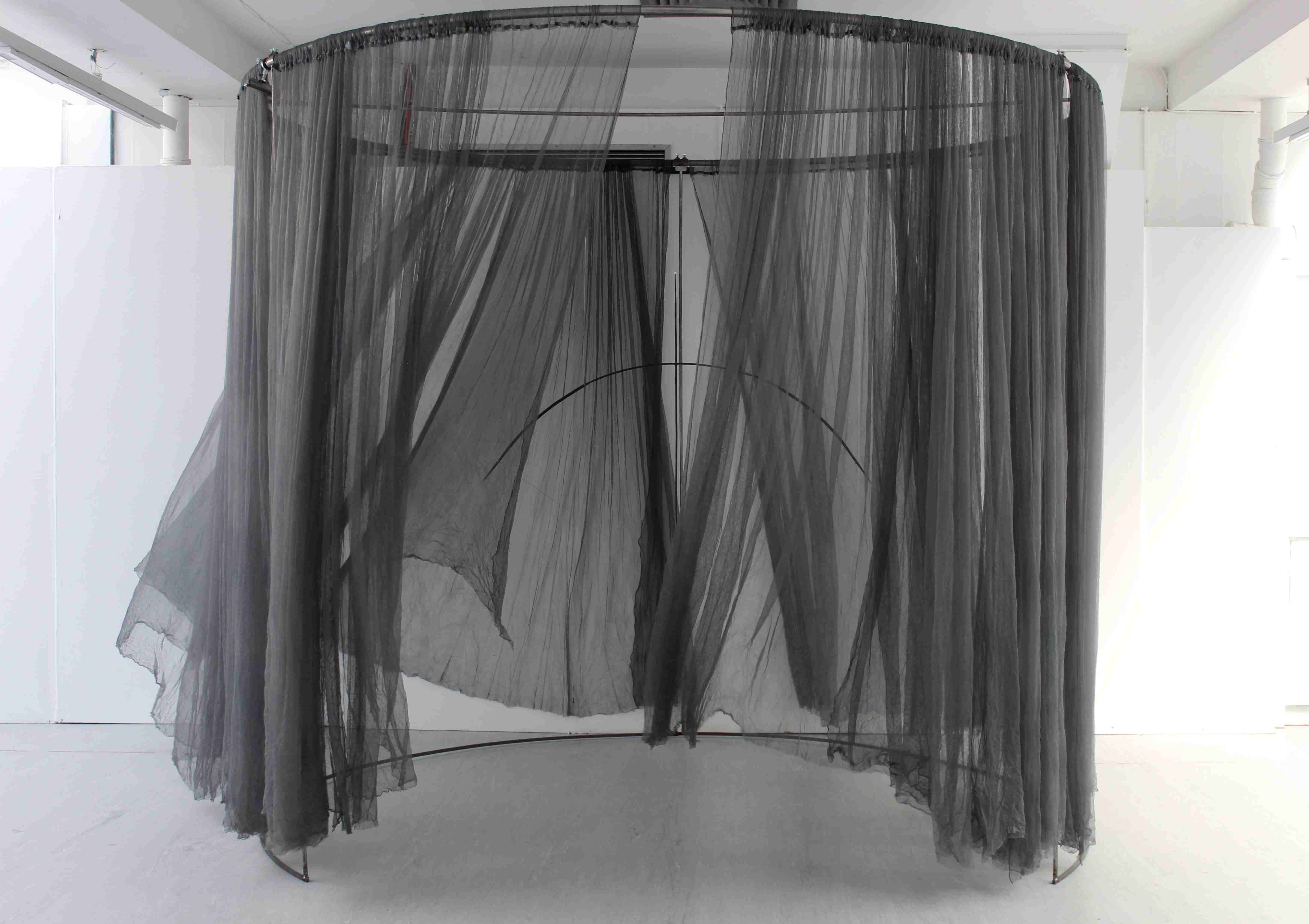 Large instillation made of a steel hoop suspending a grey veil that moves with the airflow. Inside the structure is a suspended bow and arrow, with a pearl sitting on the tip of the arrow.