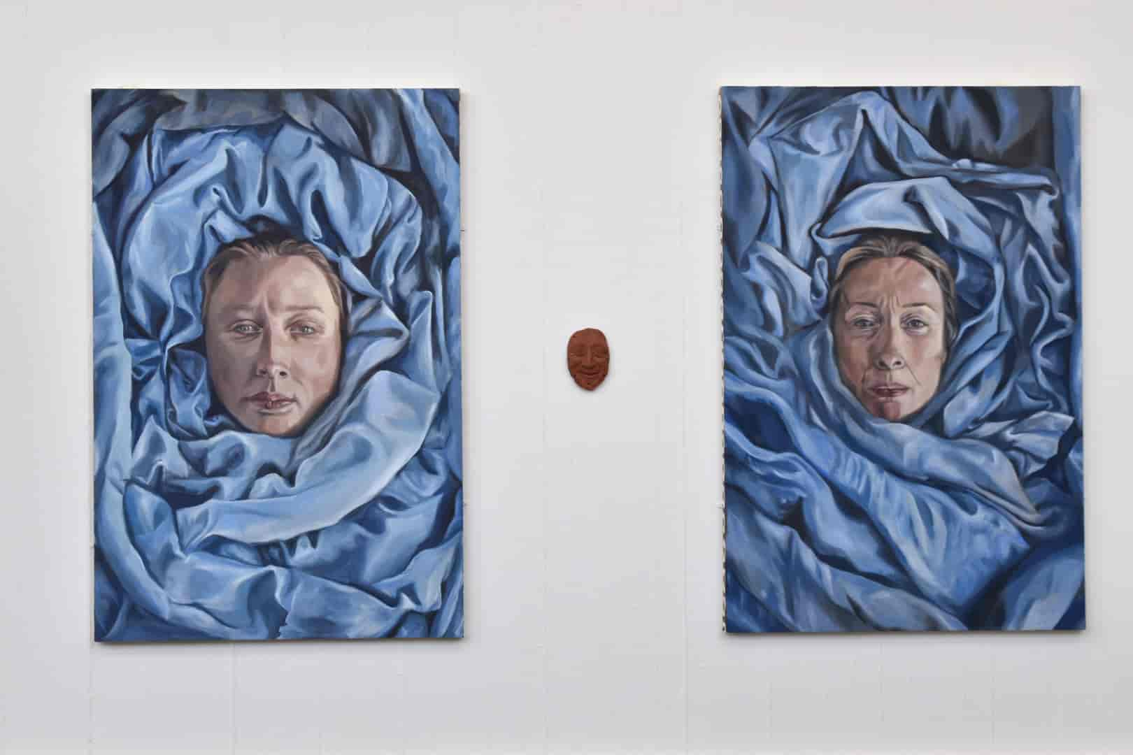 An image displaying two paintings separated by a ceramic sculpture on a white wall. In each painting there is a woman's portrait, wrapped in blue material. the sculpture in the middle is a man's portrait/mask in a terracotta clay..