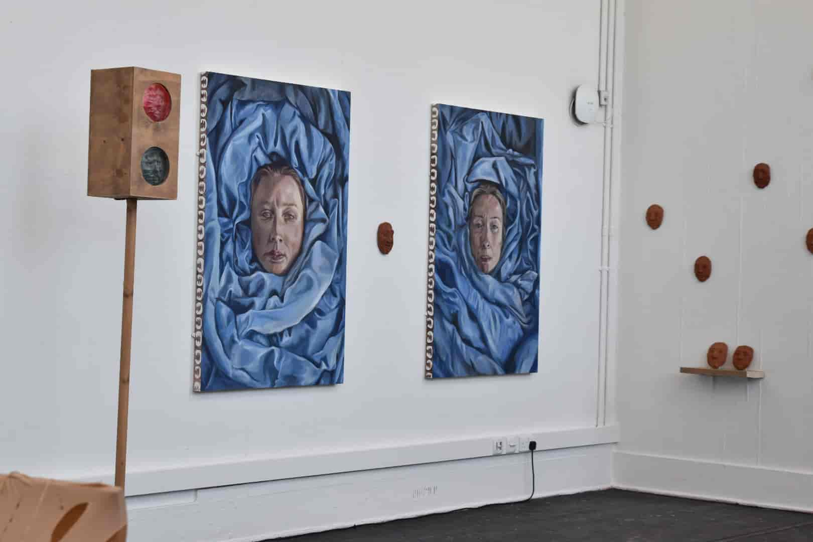 An image displaying a white walled gallery with two paintings and ceramic sculpture's mounted on the wall, as well as a wooden traffic light sculpture stood in front of the work.