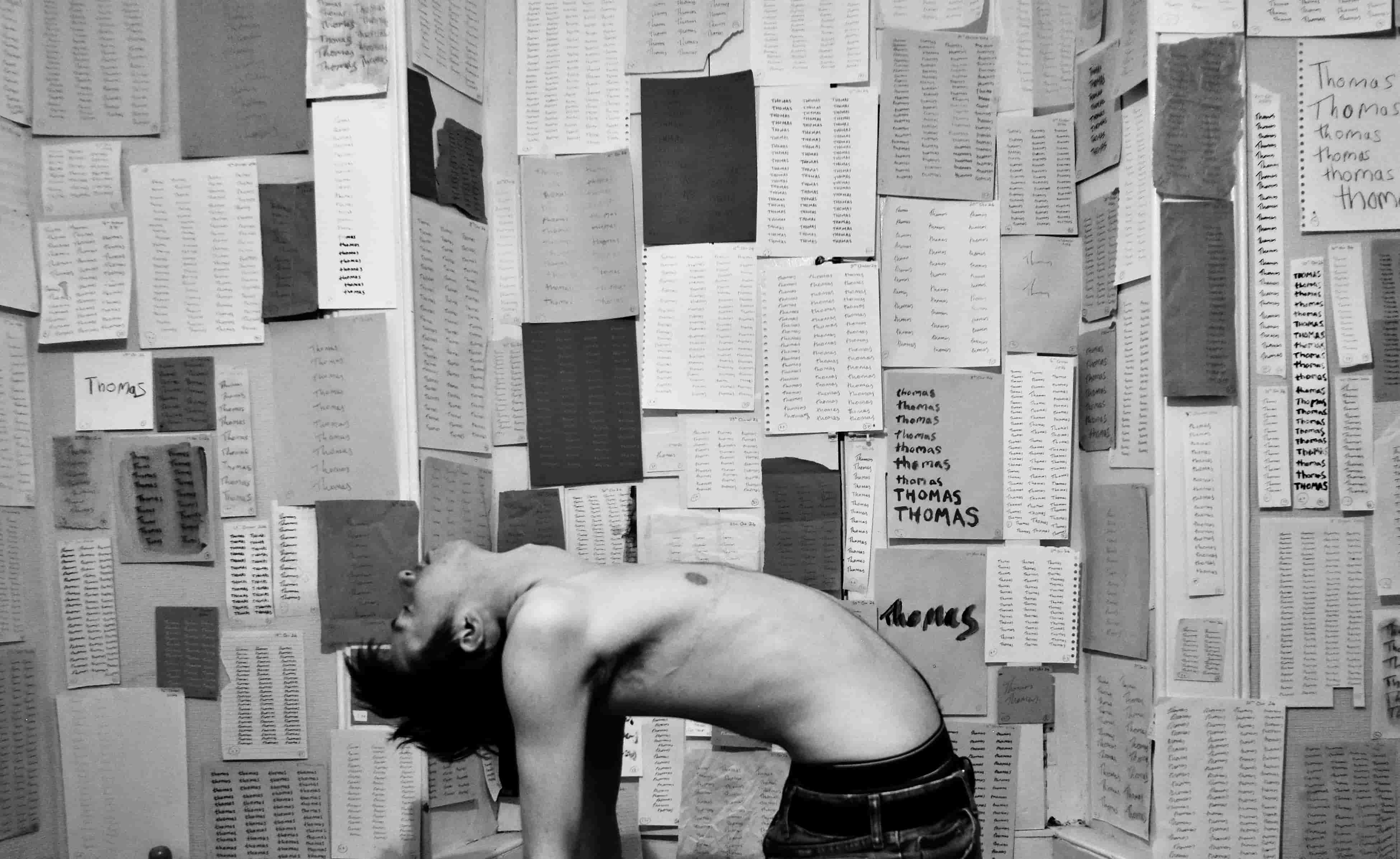 A photo of a man bending backwards with his shirt off in front of hundreds of papers on a wall, all of which read 'Thomas' repeatedly