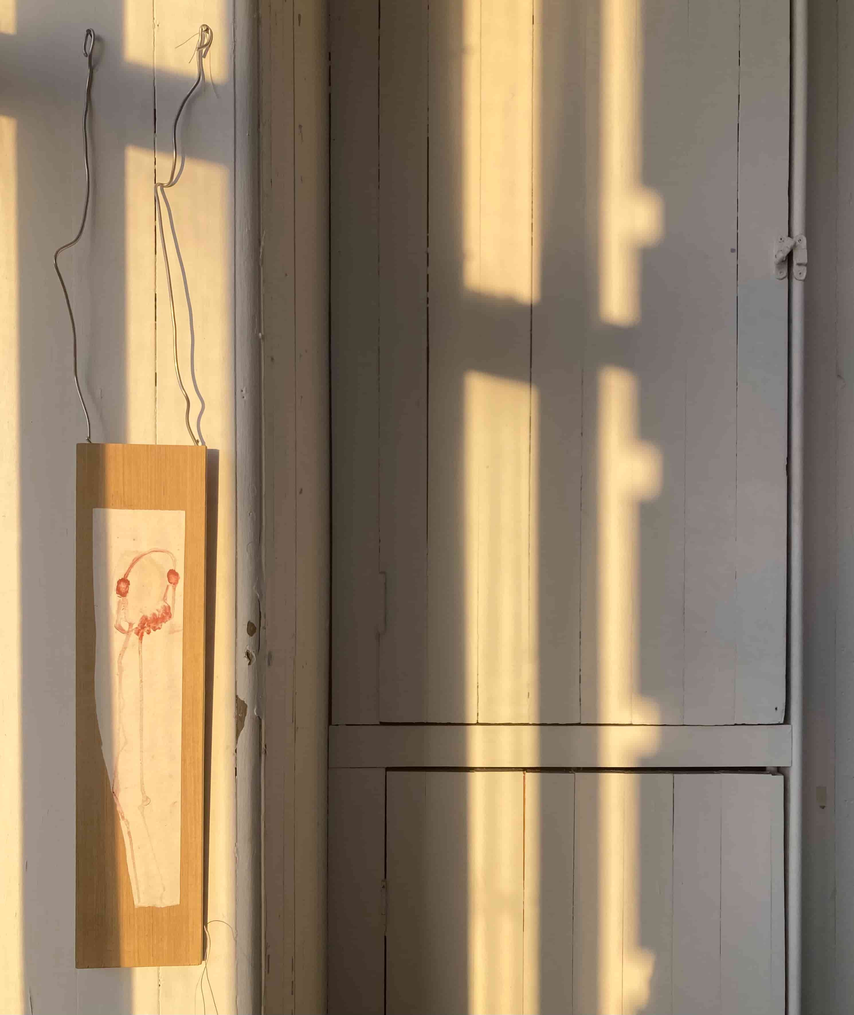 A piece of wood, hung by wire and with a painting of a head on legs on it, is suspended against a sunny wall