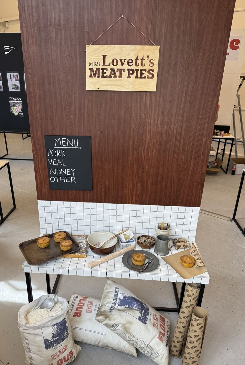 a full view of the display stand with Mrs Lovett's Meat Pies sign and menu on wooden texture background with tiled table showing graphic props (flour bags, lard and pie packaging) in situ with pies and various baking utensils