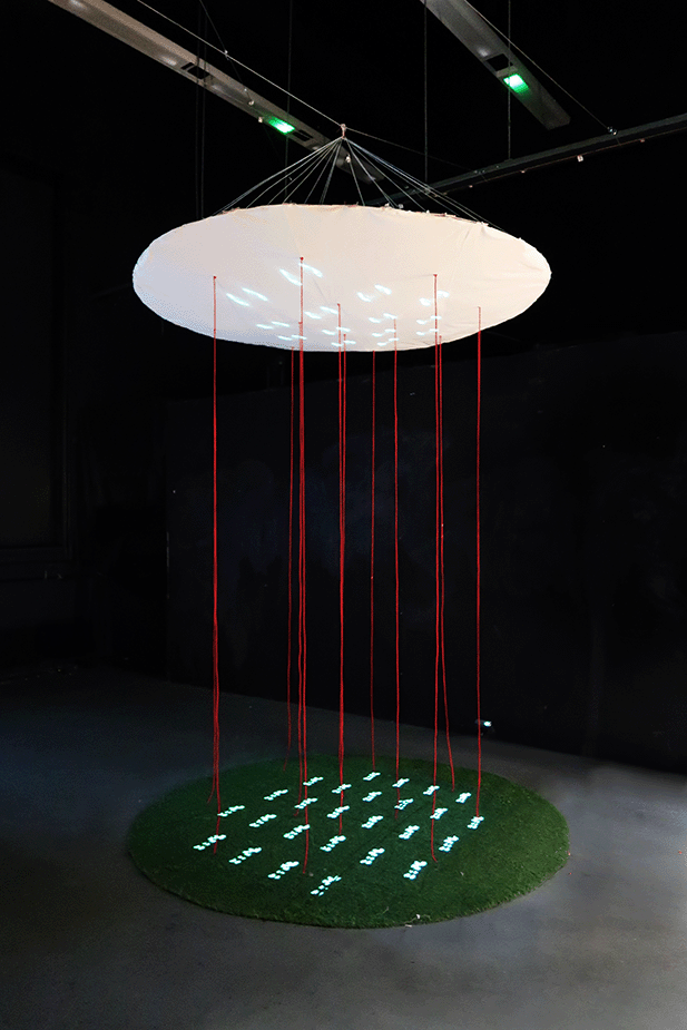 Circular installation with red strings connecting a glowing canopy and a grassy base displaying small human figures.