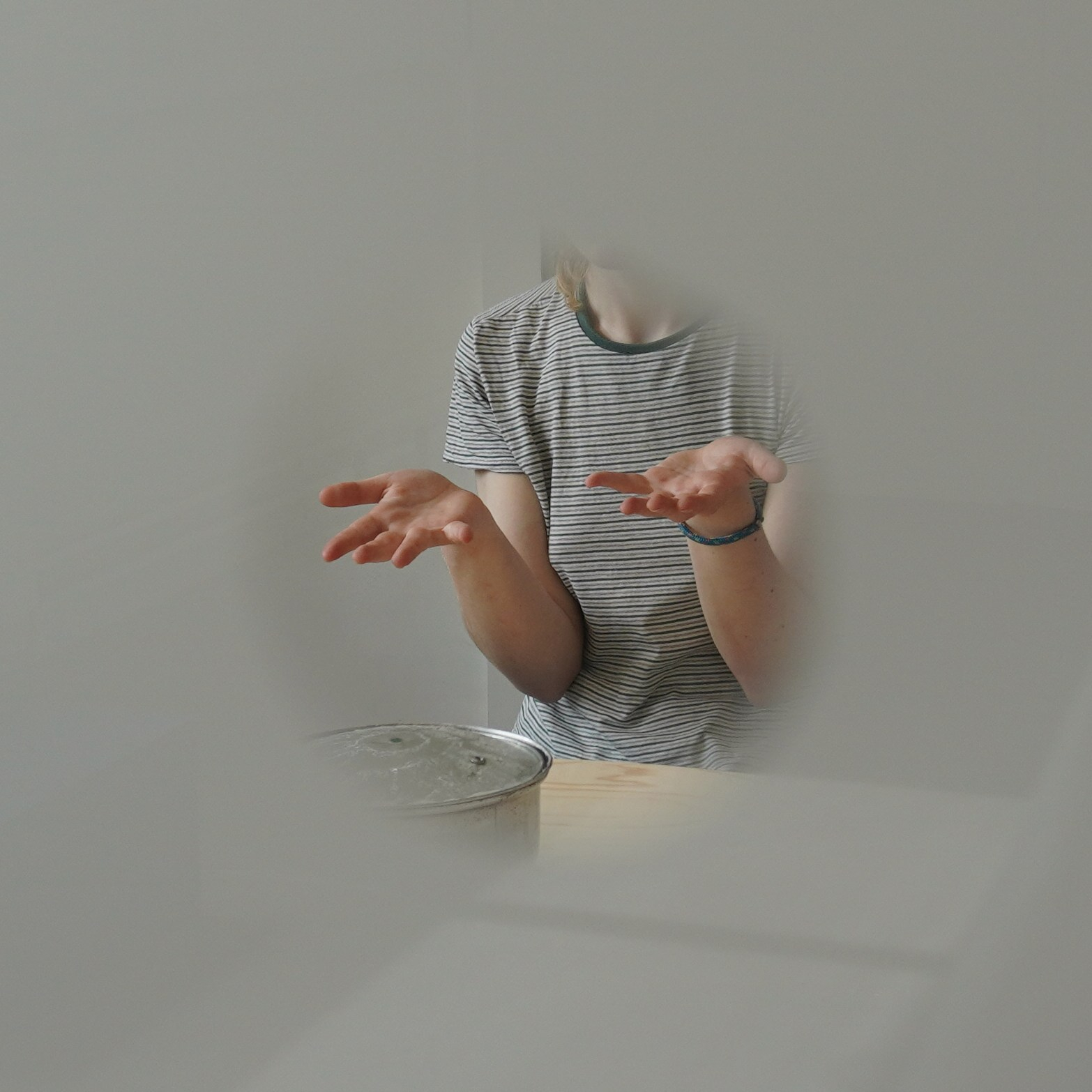 A person participating in the multisensory participatory work, 'I'm just here to absooorb.', captured through the hole on the windows. The person is opening up their palms of both hands in front of them, sitting around the table, facing the pot.