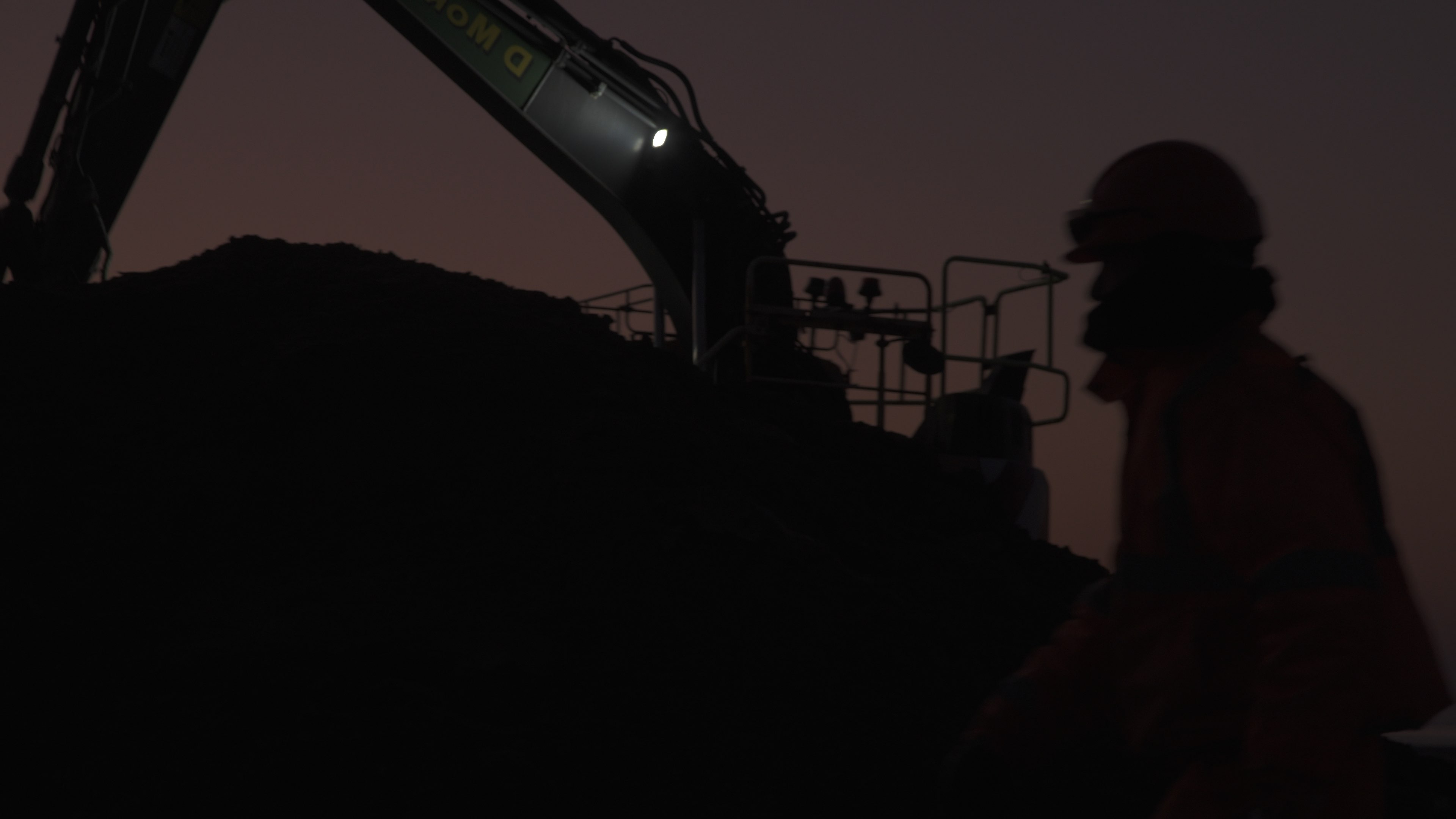 Silhouette of construction worker walking in front of a sunrise. Behind him there is a silhouette of a digger with a bright light emitting from its arm.