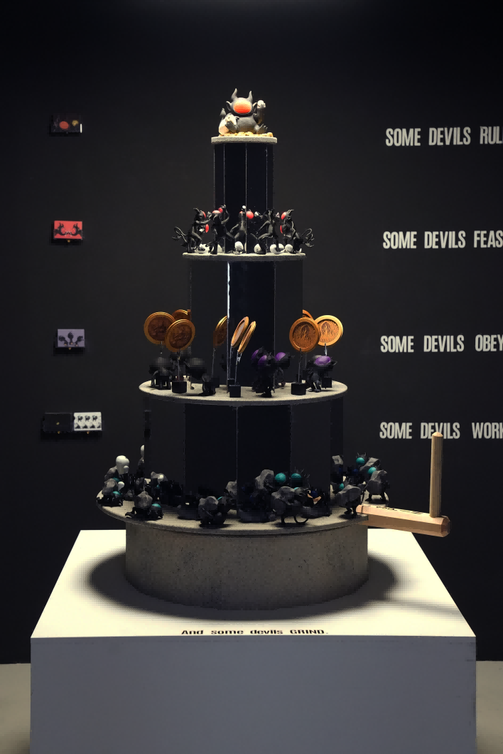 A tiered praxinoscope-inspired sculptural installation stands at the center of a dark exhibition space, viewed through an open doorway. The rotating tower features layers of silhouetted devil figures engaged in labor, echoing themes of capitalist hierarchy. On the black walls behind, white text reads “Some Devils” in repeated lines, reinforcing the work’s title and critical message.