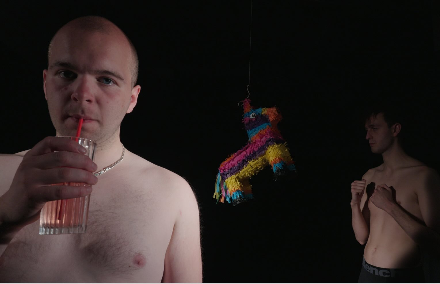 a man sips juice while another prepares to hit a pinata with his fists 