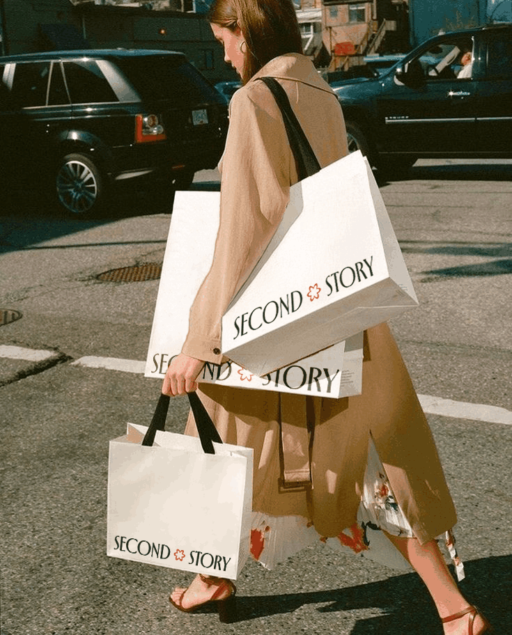     Person walking across a street carrying large white Second Story shopping bags with black handles.