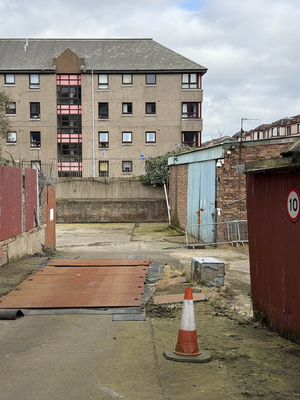 A back garden covered in screed and surrounded with fences. Residential buildings tower over site