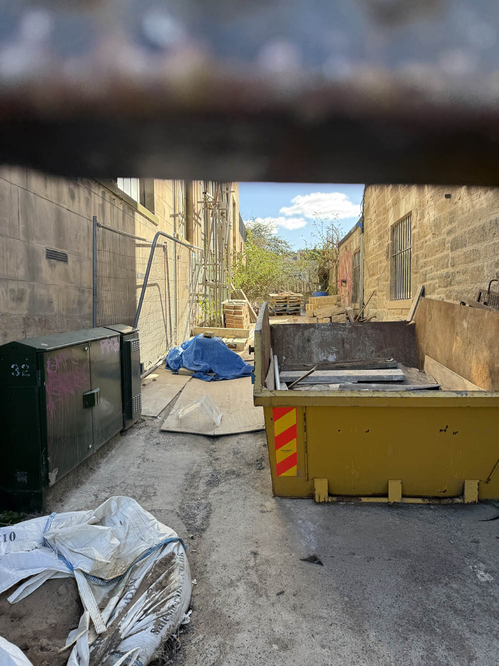 Alley between two building is closed off with a gate, skip and clutter are visible