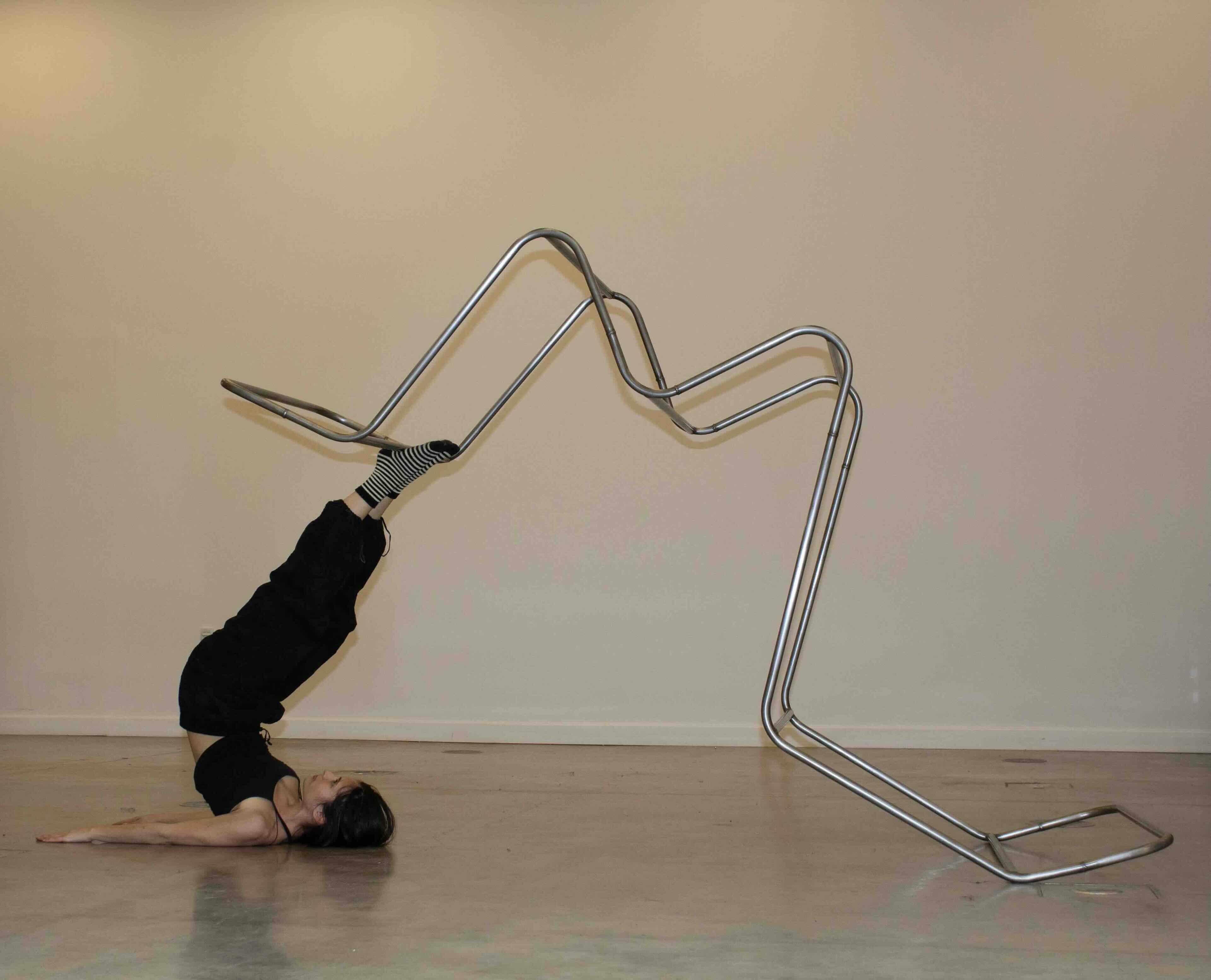 A person lies on their back in an empty room with their feet and legs in the air, lifting up a metal frame made of steel pipes bent and welded into an 'm' shape.