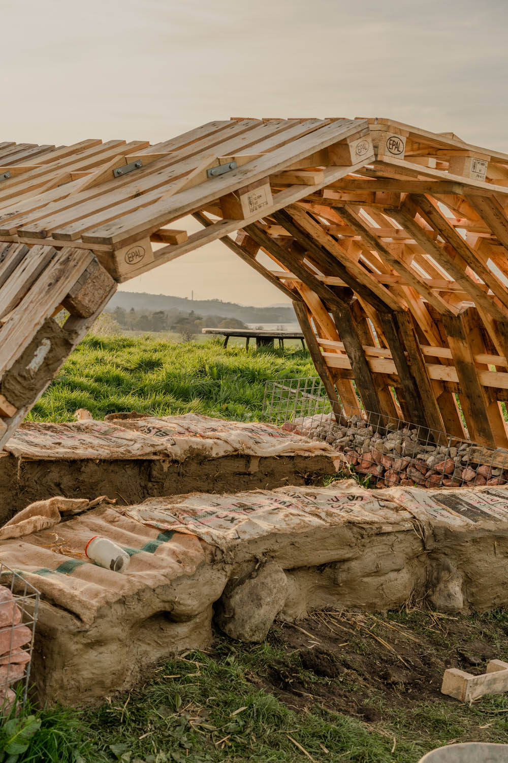 Photo of the pavilion covered with arched roof made of three rows of pallets covering two rows of benches; time of the day is sunset