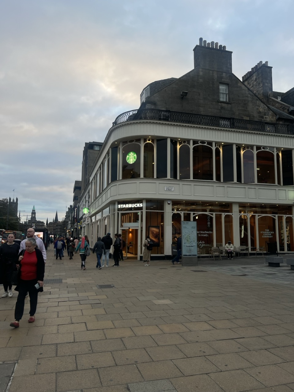 A street-level photograph of the existing Princes Street corner site in Edinburgh, showing the curved listed building façade, surrounding pedestrian activity, and views towards the city skyline within the busy urban context.