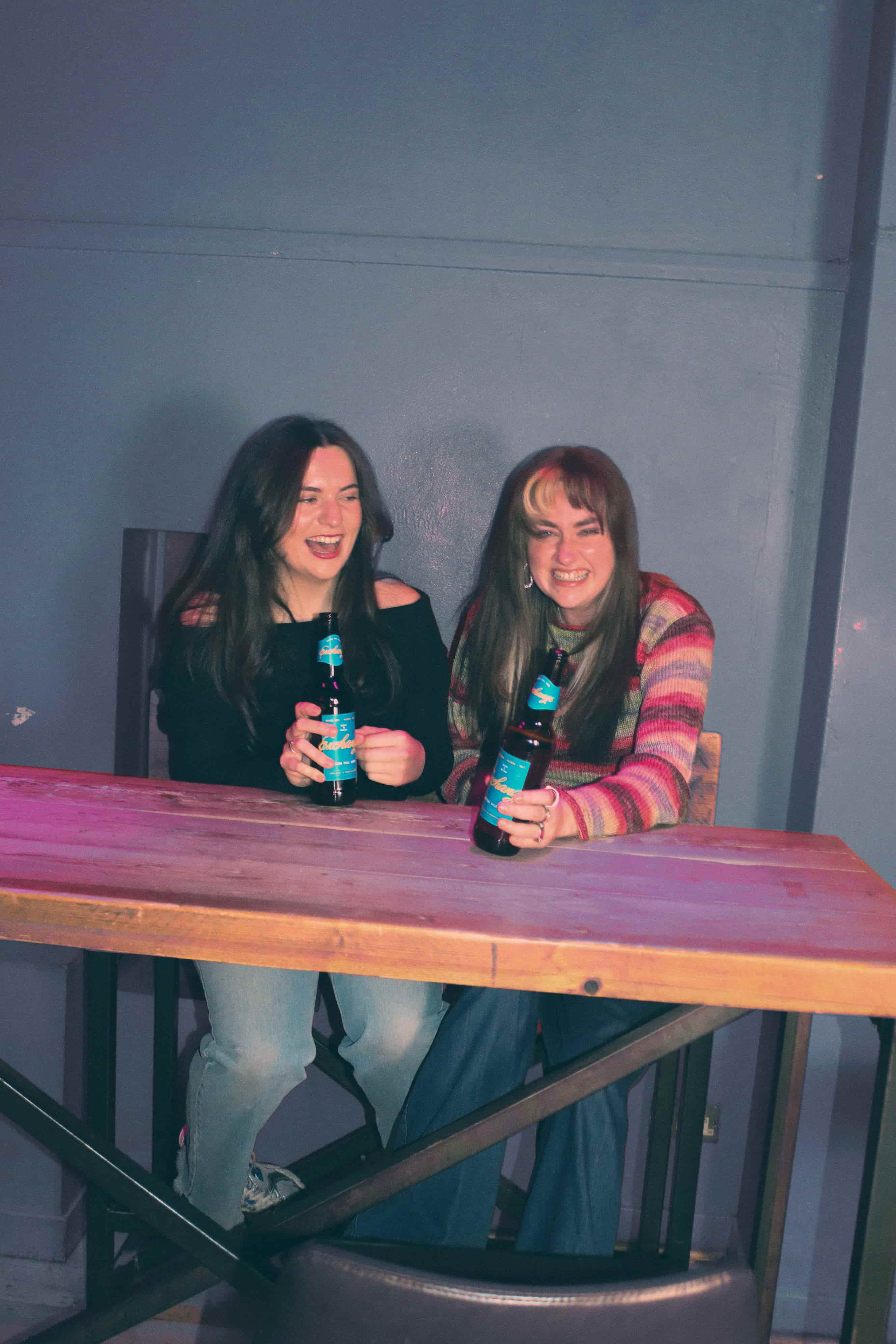     Two people sitting at a wooden table holding ‘Exchange’ beer bottles and laughing.