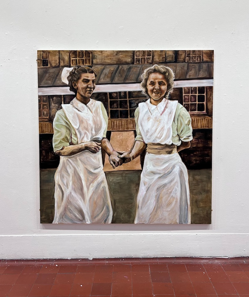 Two Nurses stnad in front of a building. They are in green uniforms. The right nurse, stands smiling for a photograph. She holds the hand of the left nurse, who appears shy. 