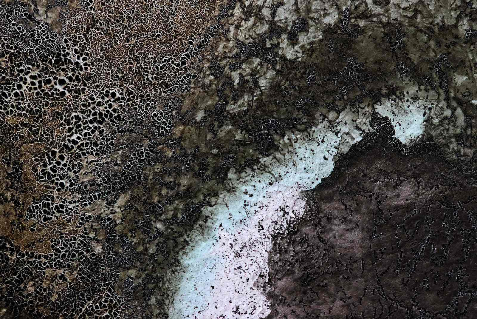 Macro photograph of natural ink on a painted surface. The image shows a close-up of dark brown, black, grey, and pale blue areas where the ink has dried into layered textures. On the left, a dense network of pale crackled lines forms a web-like pattern over a dark background. Across the centre and right, darker sedimented areas meet lighter blue-white patches, with granular specks, branching marks, and uneven tide lines revealing where the ink has pooled, separated, and dried.