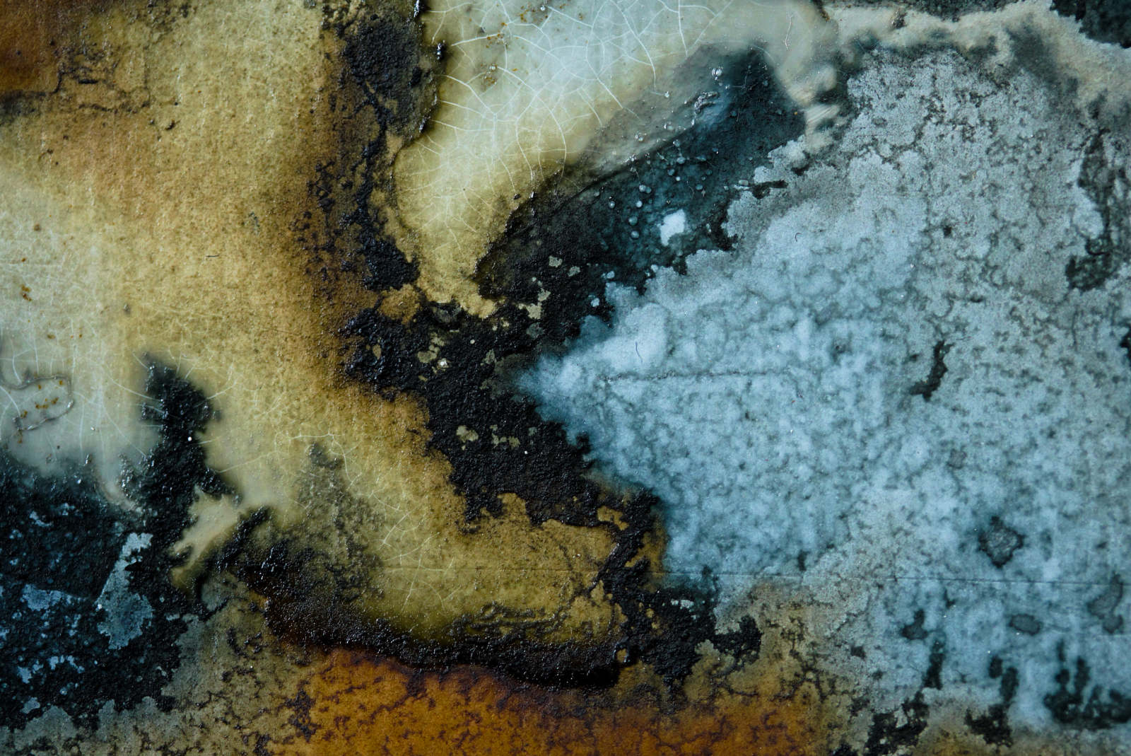 Macro photograph of natural ink on a painted surface. The image shows a close-up of blue, turquoise, ochre, cream, brown, and black areas where the ink has pooled and dried. A large pale blue section fills the right side, bordered by dark branching stains and granular pigment. On the left and centre, ochre and cream areas are crossed by black sedimented edges, fine pale crackle lines, and uneven tide marks, showing the ink separating, settling, and crystallising across the surface.