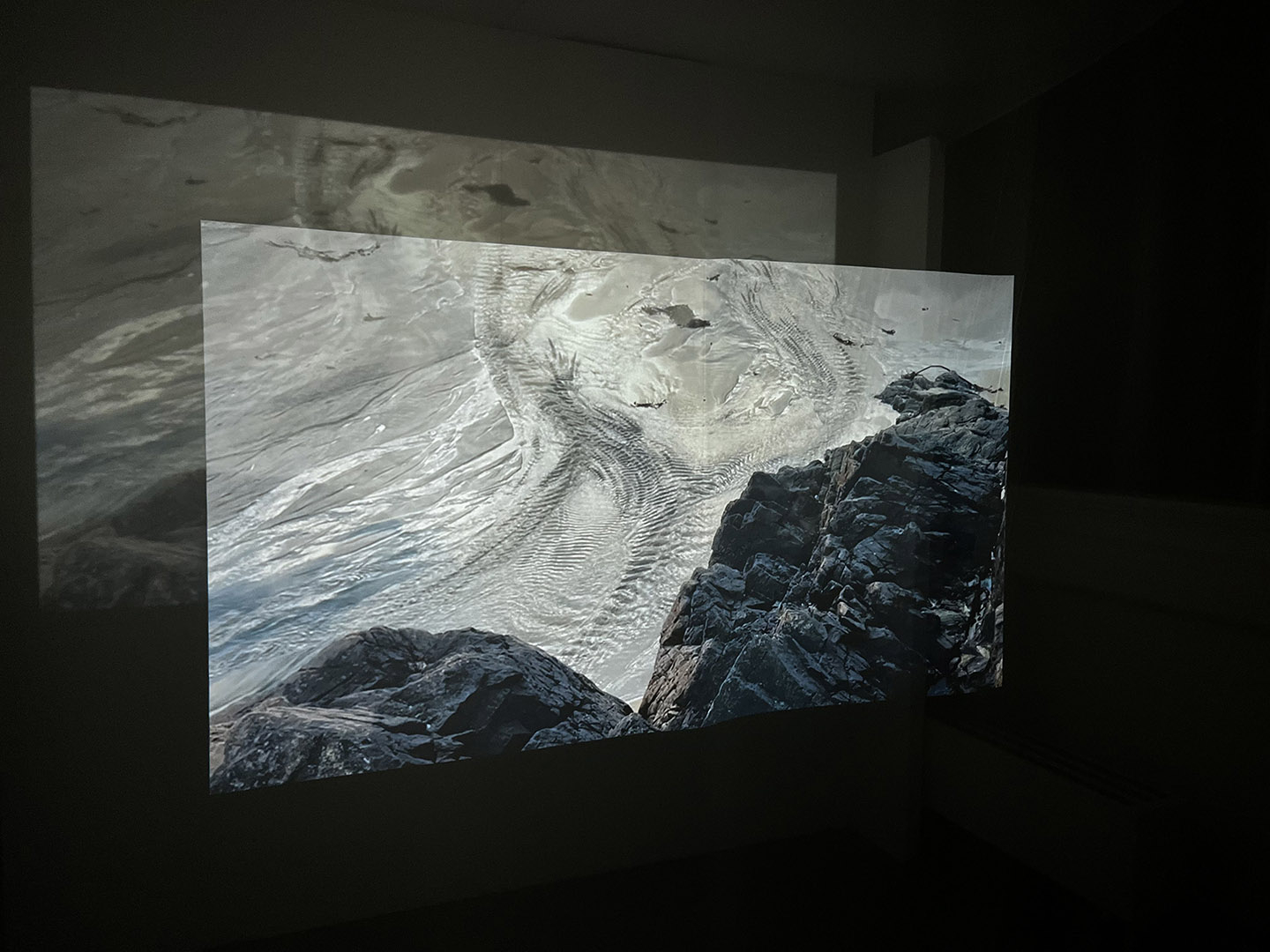 Photograph of projected film in dark room showing the ripples of beach near rocks..
