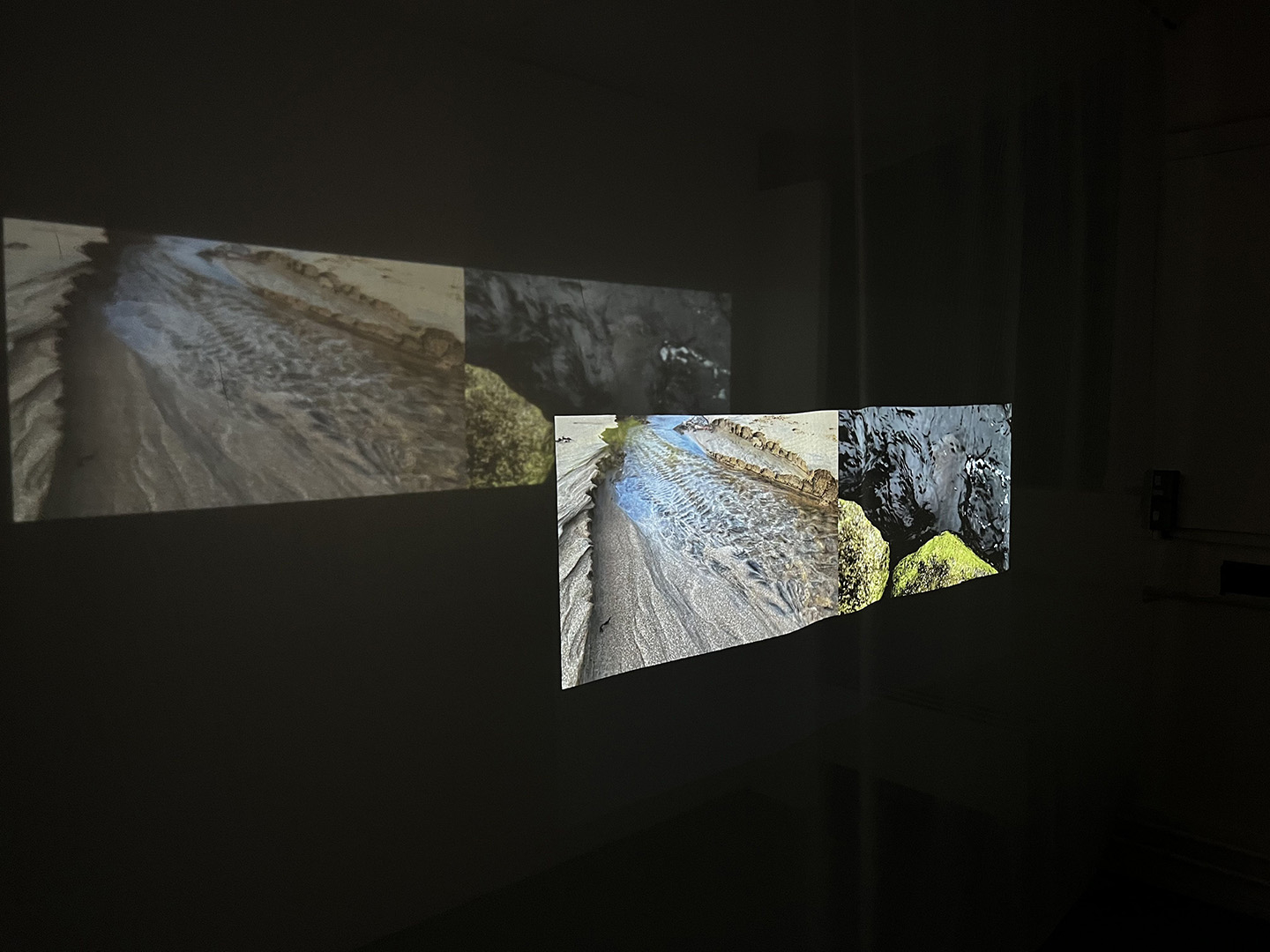 Photograph of projected film in dark room showing a beach river and a peat river with green, mossy stones..