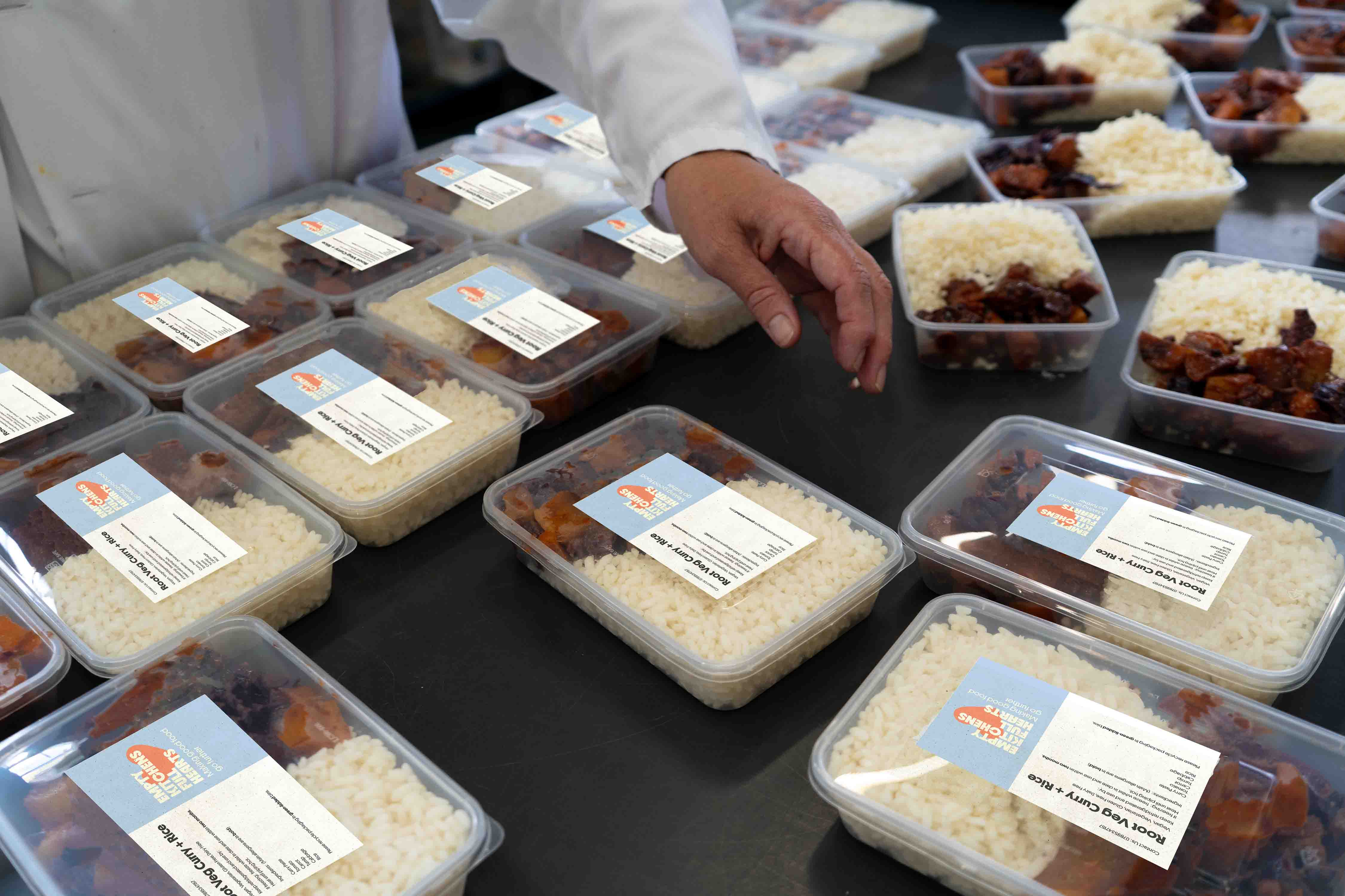 Tupperwares full of meals produced by the charity with designed informational labels describing the meal 'Root Veg Curry and Rice' alongside ingredients and allergens ready to be packed